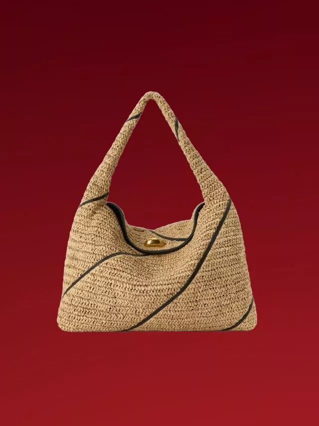 Woven straw hobo handbag with black trim and gold clasp, studio product photo on deep red background