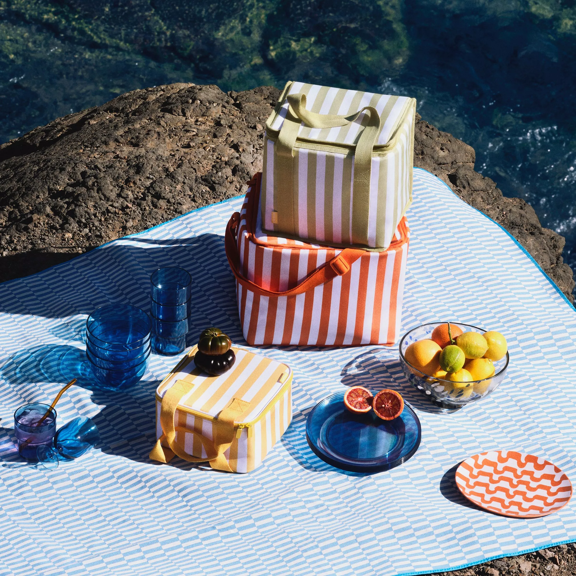 Striped cooler bags and picnic set on blue mat by rocky coast, with citrus fruit, plates and blue glasses.