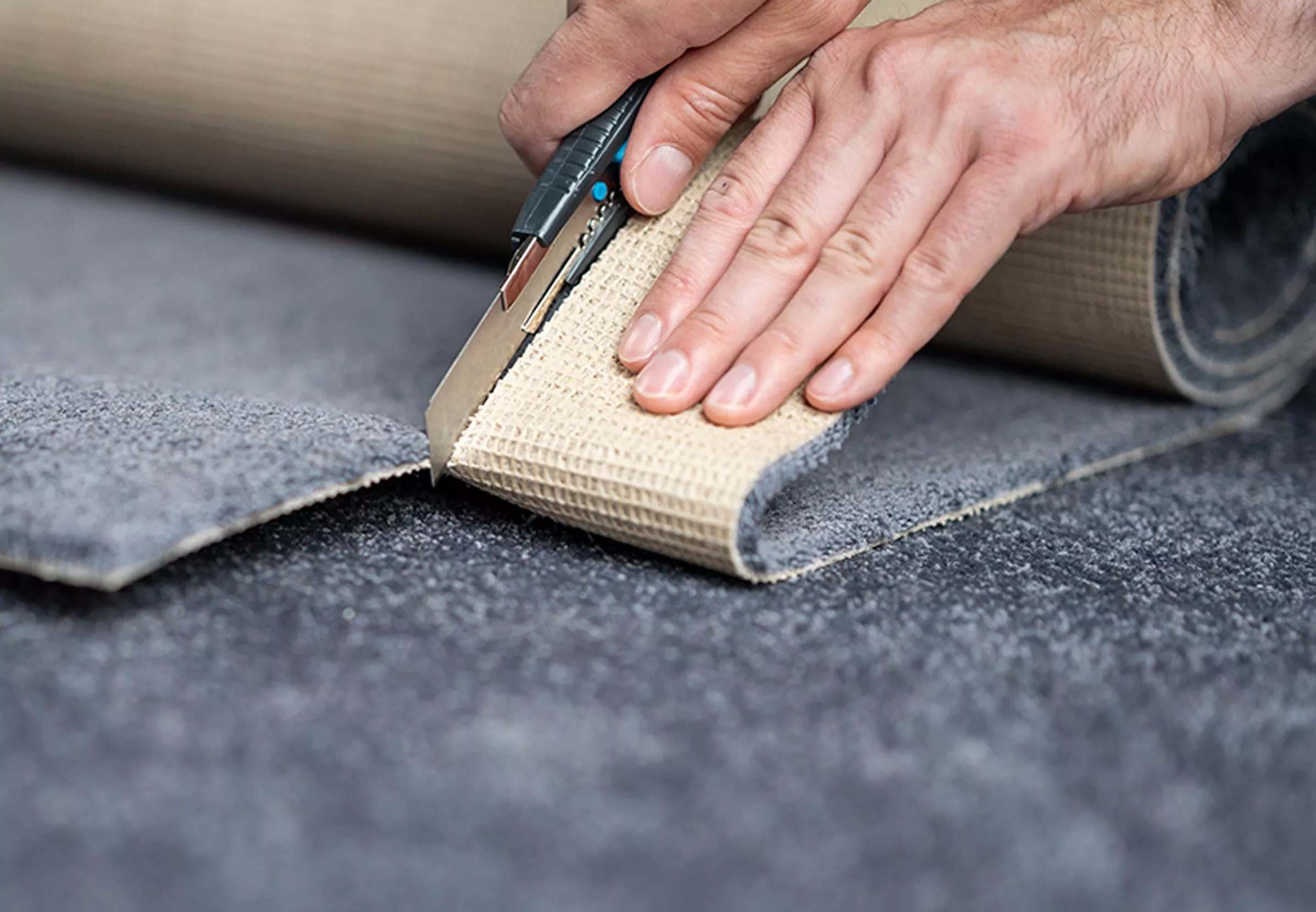 Hands cutting grey carpet with a utility knife on a textured surface. Carpet installation process.