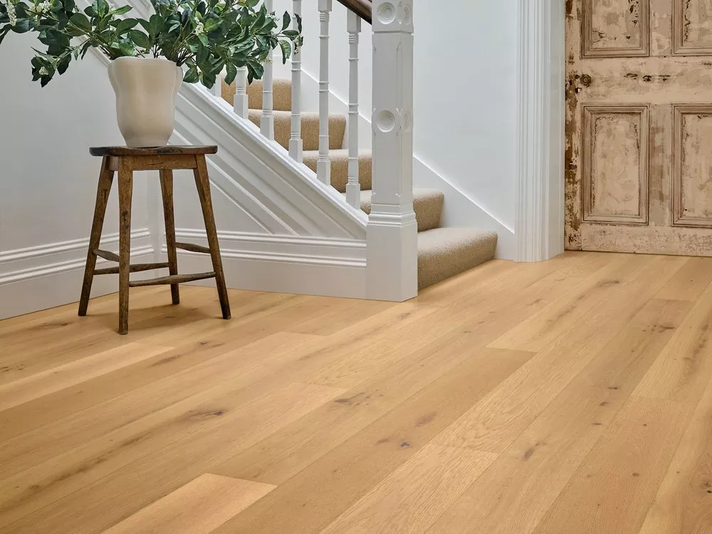 Light oak wooden flooring in a bright hallway with staircase, rustic door, and plant on stool.