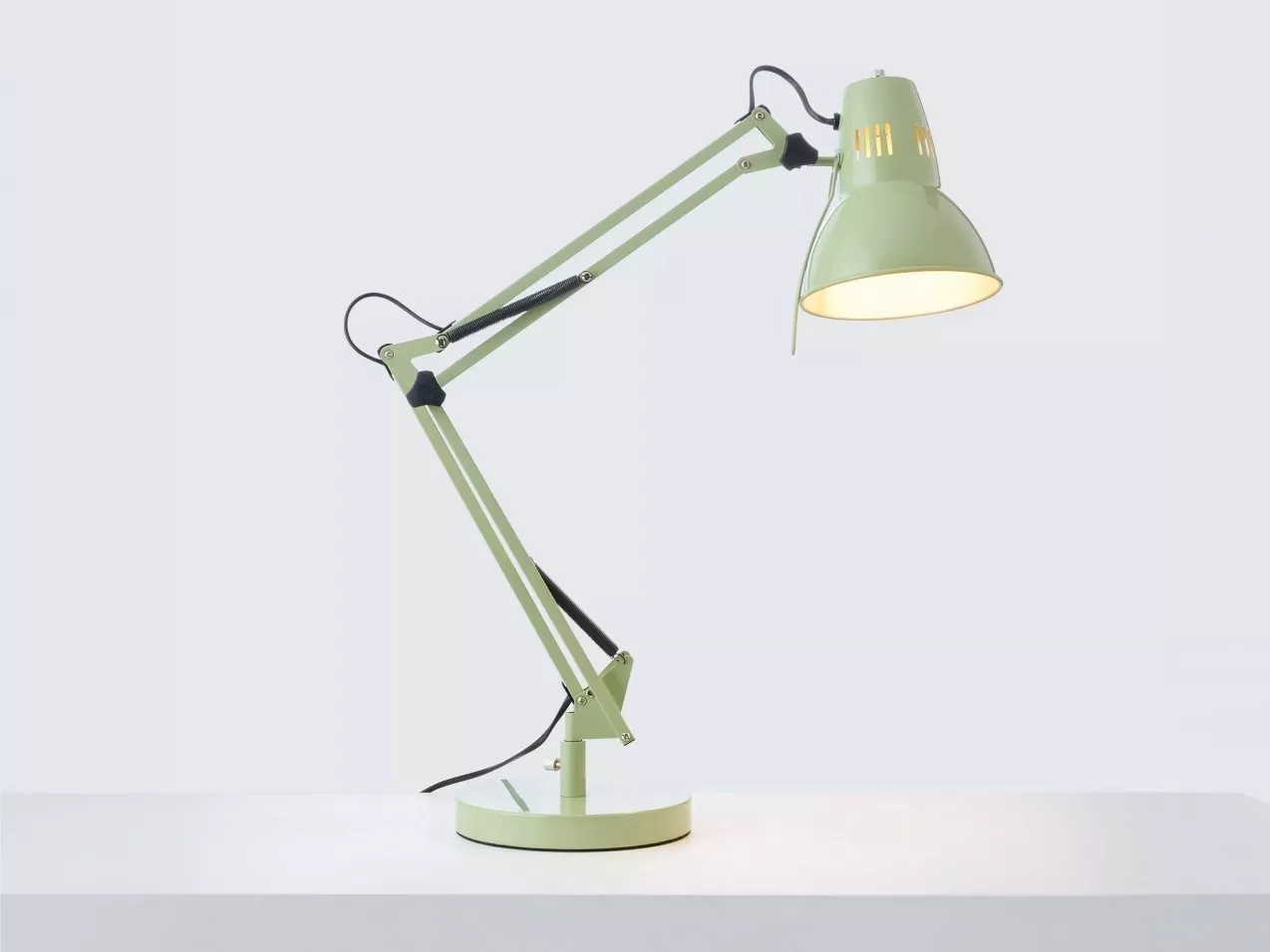 Pale green adjustable anglepoise desk lamp with metal shade, lit bulb, and articulated arm on white background
