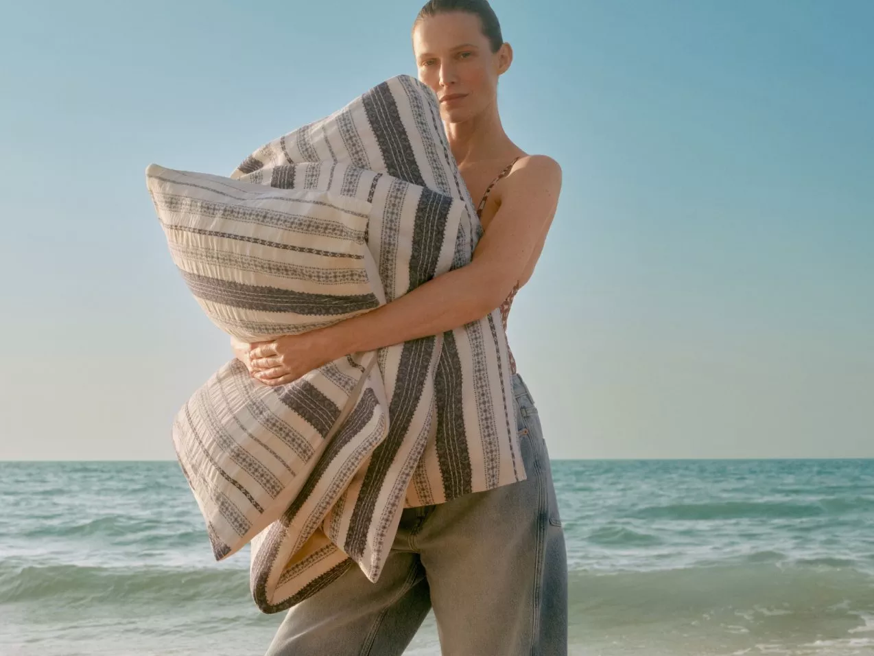 Woman holding large striped cushion by the sea, outdoor lifestyle photo showcasing coastal home décor textiles