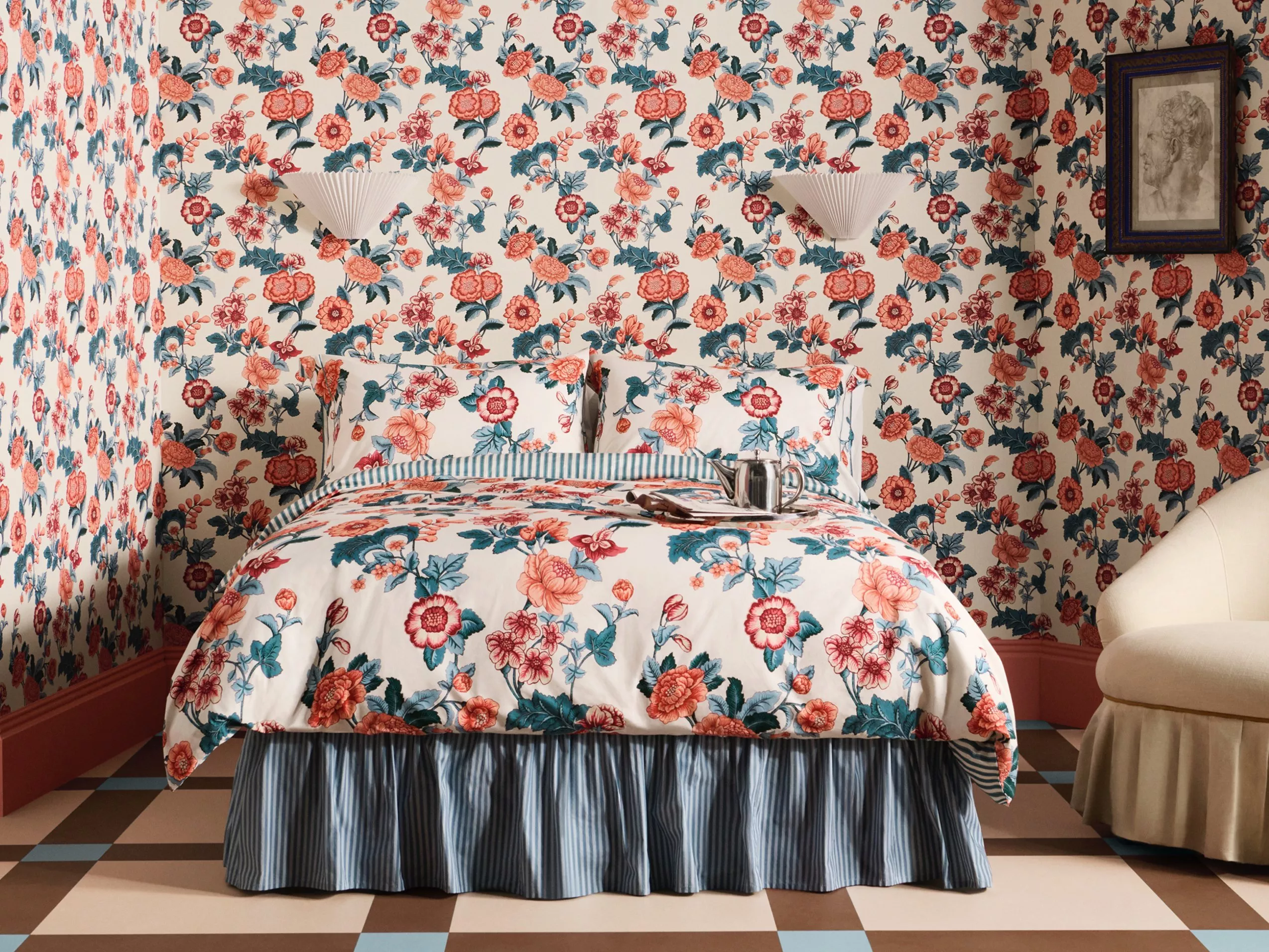 A bedroom with floral wallpaper and matching bedspread. The room features a patterned floor and cream coloured arm chair.