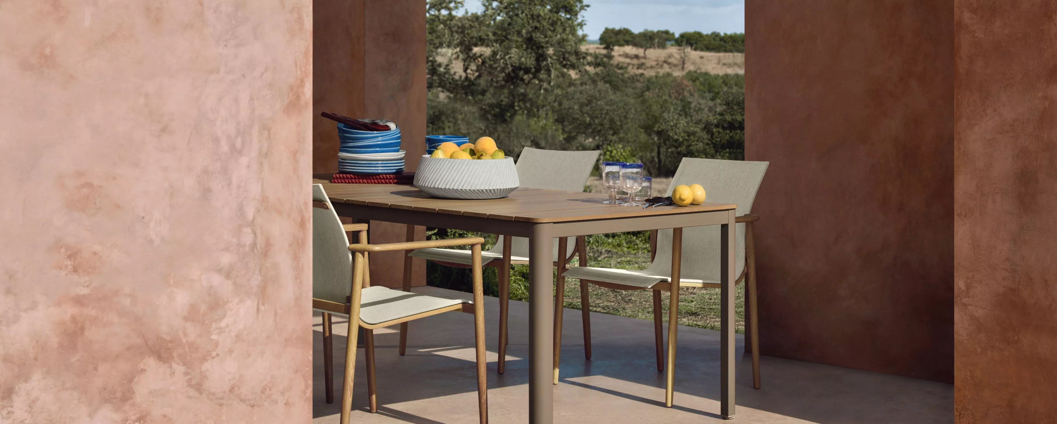 Outdoor dining table with minimalist chairs, fruit bowl, plates and glasses, framed by terracotta walls and garden view.