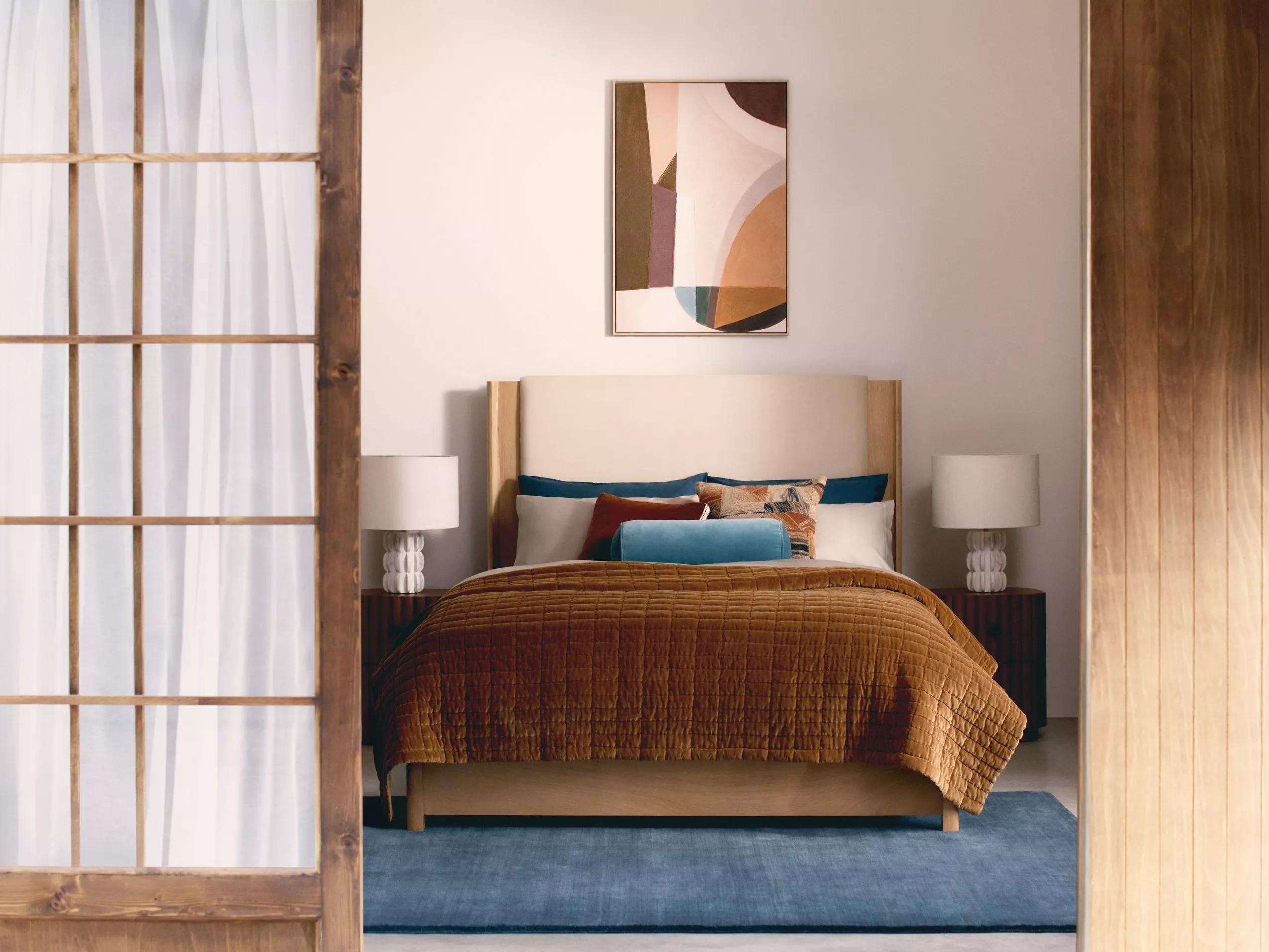 Bedroom with upholstered bed, mustard quilt, bedside lamps and abstract wall art in warm neutral decor.