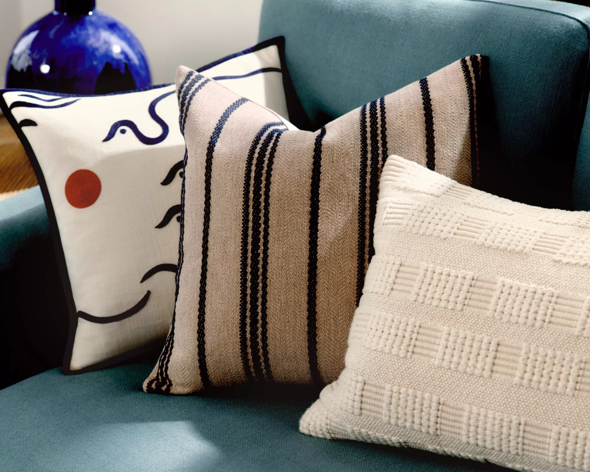 Teal sofa with modern decorative cushions, striped and textured cream pillows, and abstract face cushion close-up