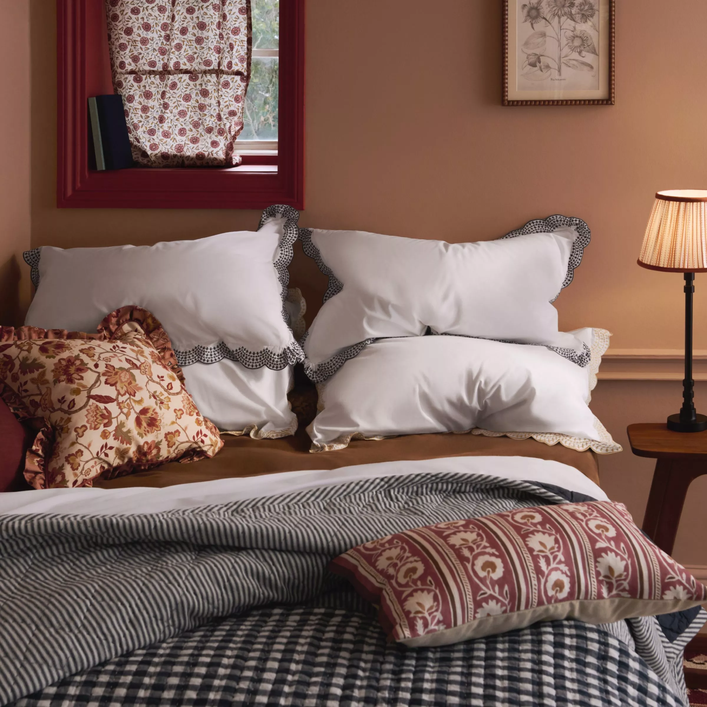 Cosy bedroom with patterned bedding, white scalloped pillows, floral curtains, framed art and bedside lamp.