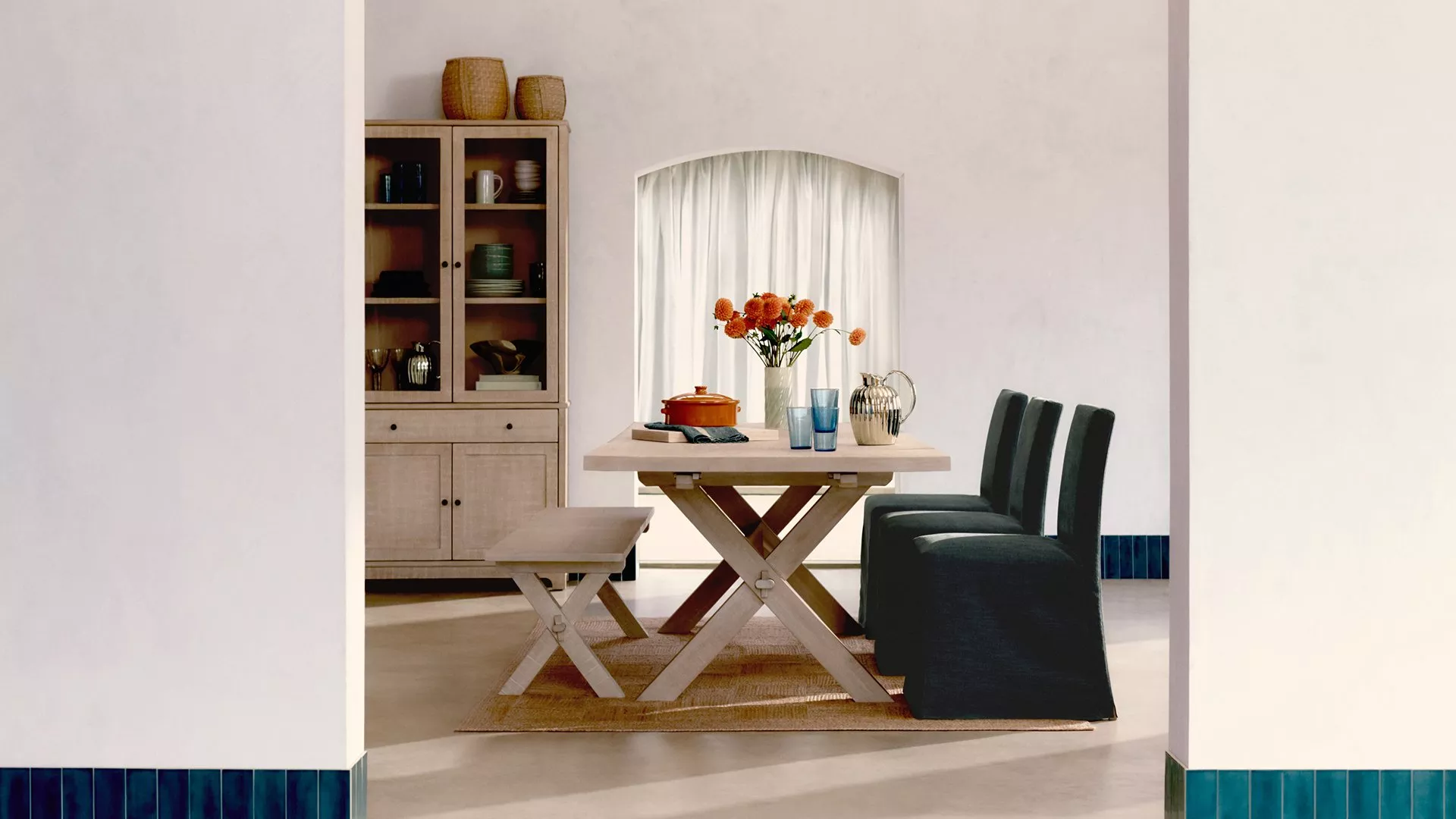 Stylish dining area with a wooden table, cushioned chairs, and orange flowers. Cabinet and decor complete the scene.