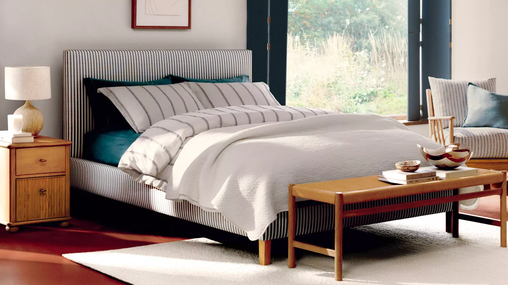 Stylish bedroom with a striped bed, wooden furniture, and soft lighting. Large window reveals a garden view.