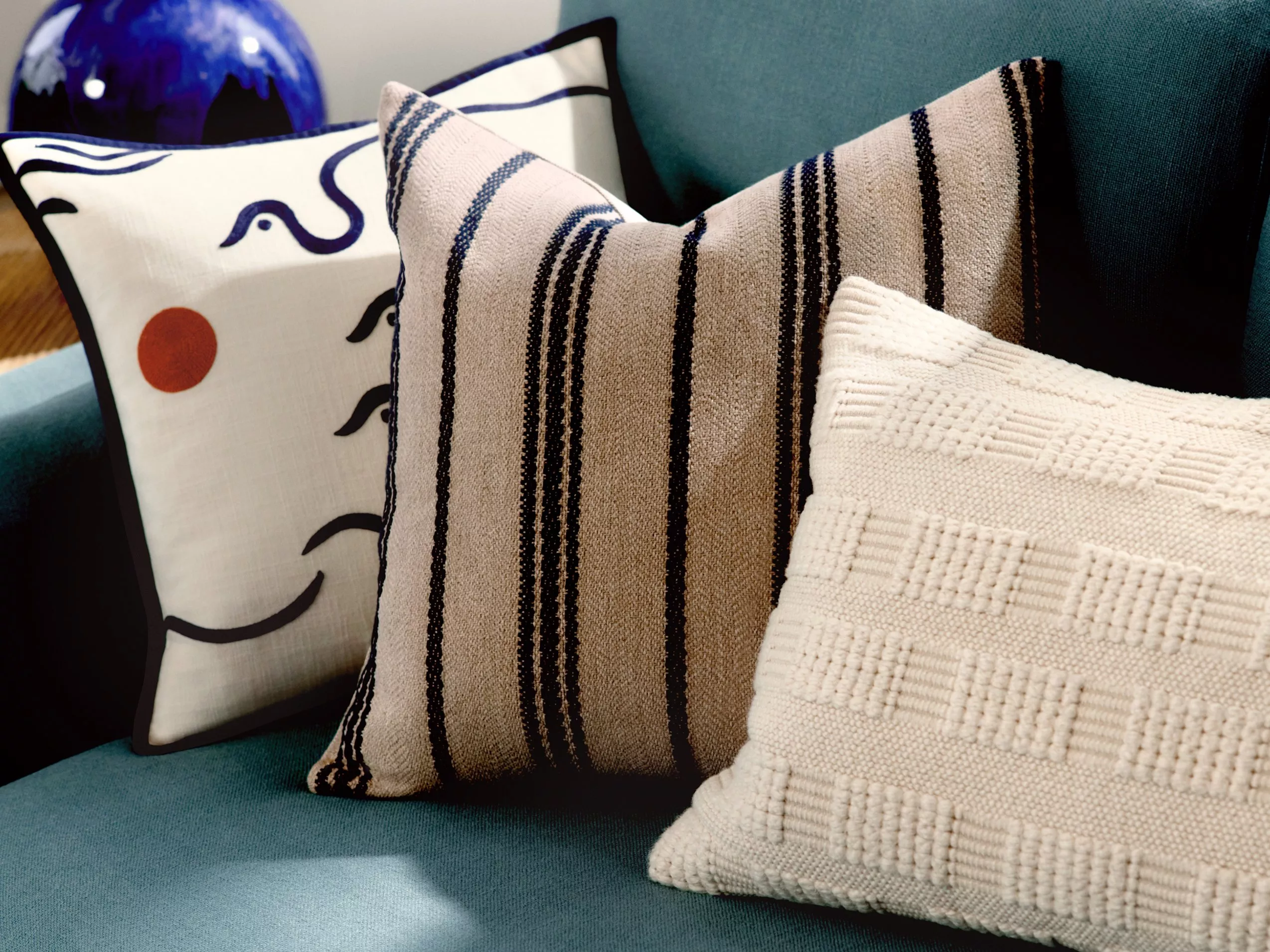 Three decorative cushions on a blue sofa: one with abstract art, one striped, and one with a textured pattern.