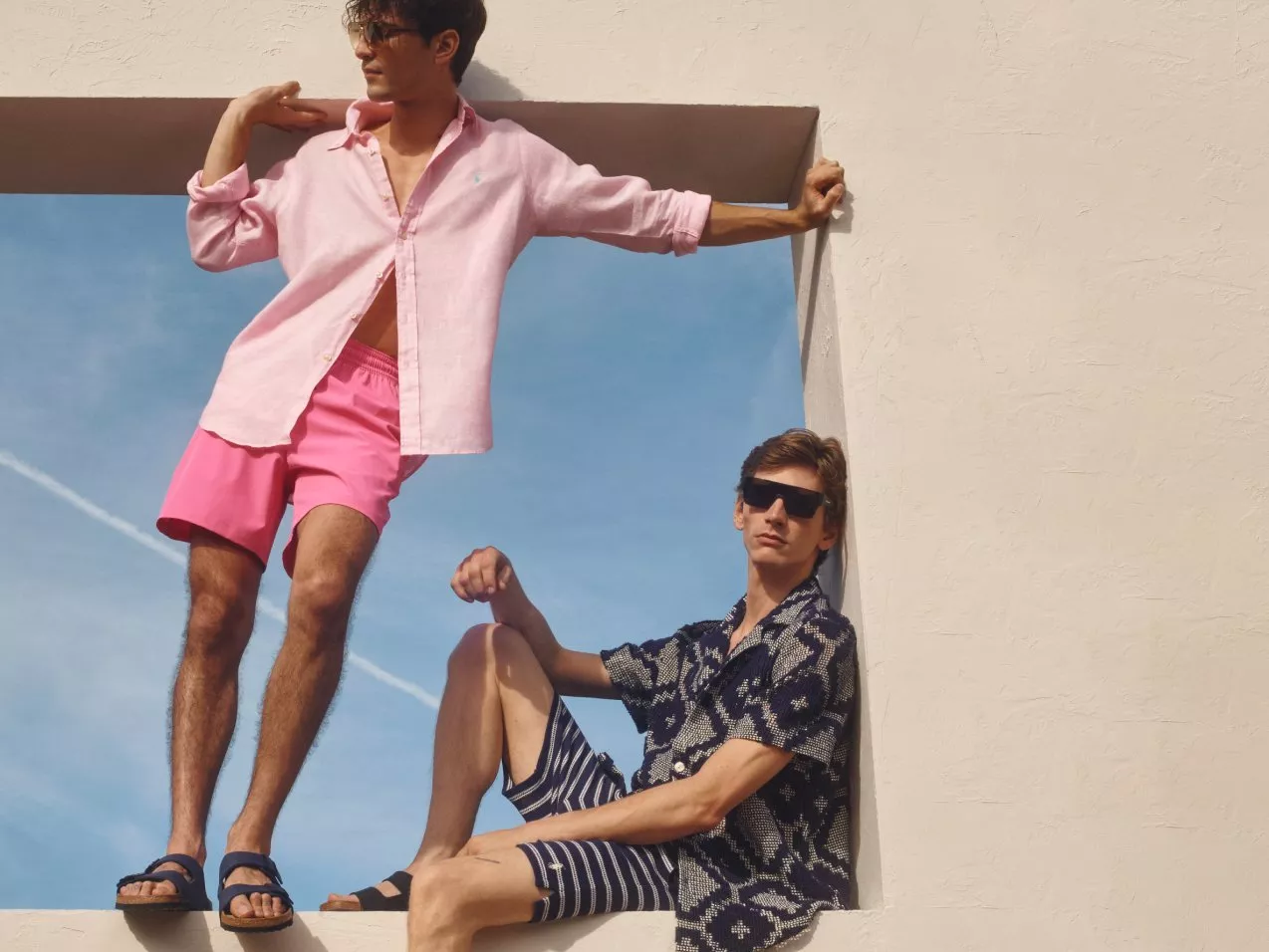 Two men in sunglasses wearing summer shirts and shorts posing by a white wall against a blue sky outdoors