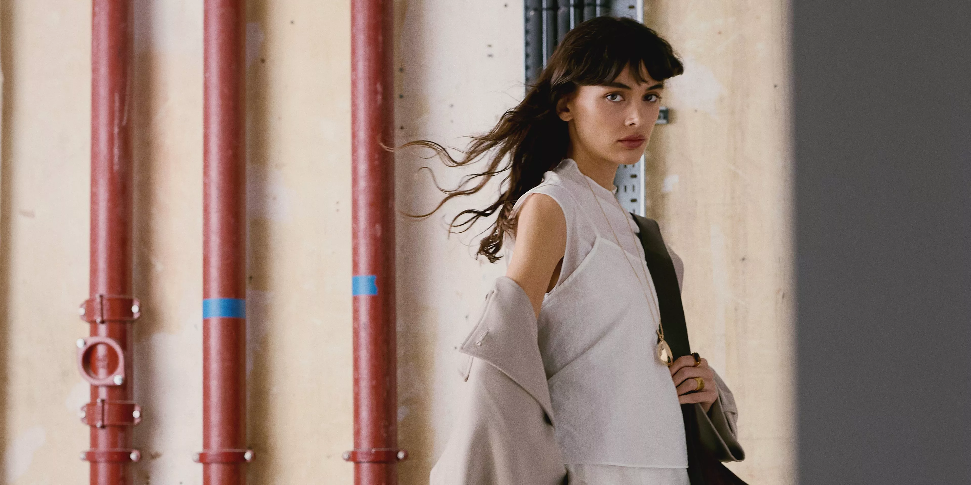 Woman in a dark, striped outfit leans against a textured, aged wall in a room with a wooden door and fireplace.