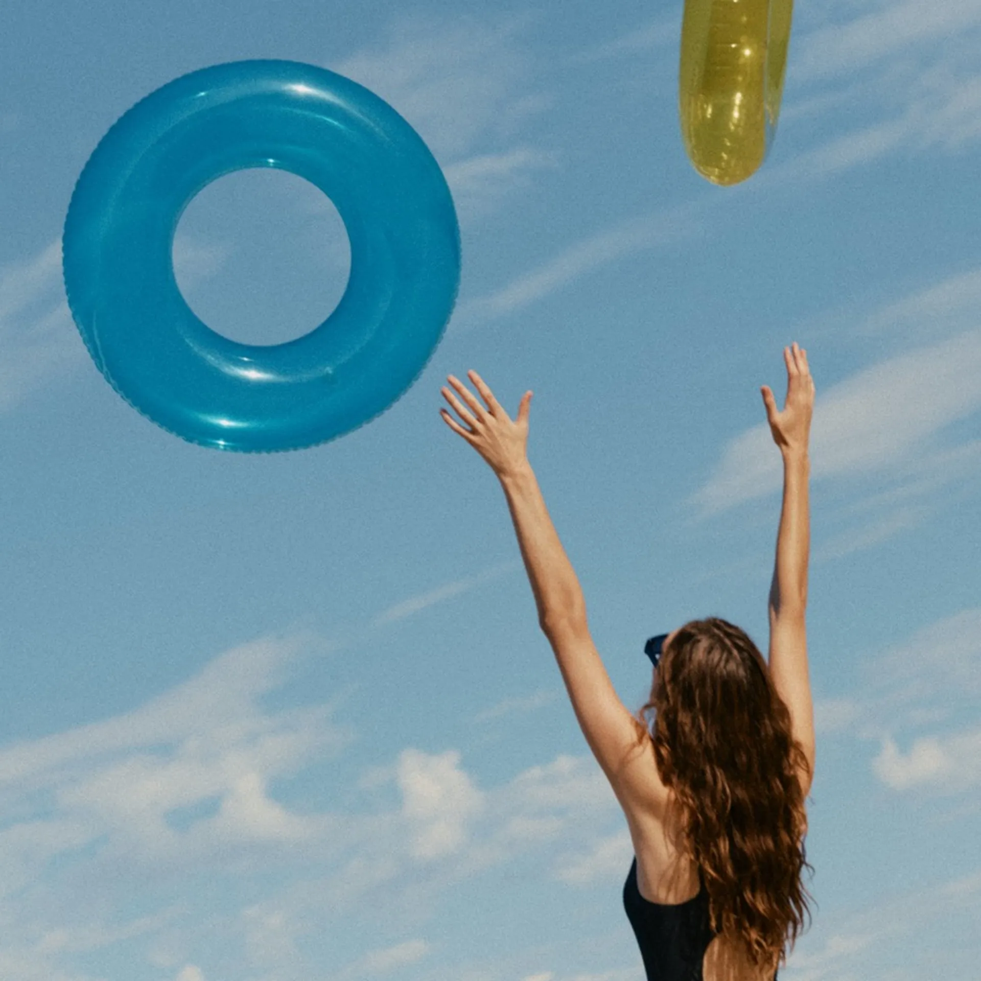 Woman in black swimsuit on sunny beach throwing inflatable rings into blue sky, summer holiday fun