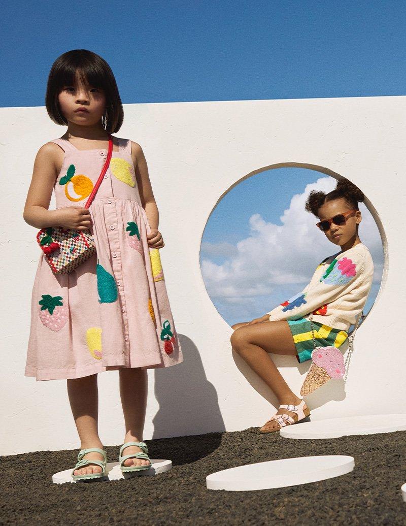 Girl standing in fruit patterned summer dress, cross body bag, and sandles, and another girl seated wearing sunglasses, fruit patterned cardigan, striped shorts and sandles.