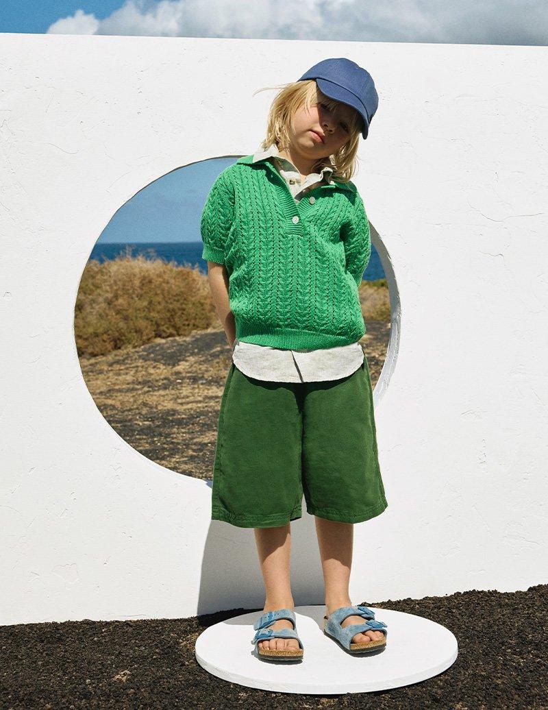 Boy in blue cap, green short sleeved sweater, green long shorts, and blue Birkinstock sandals.