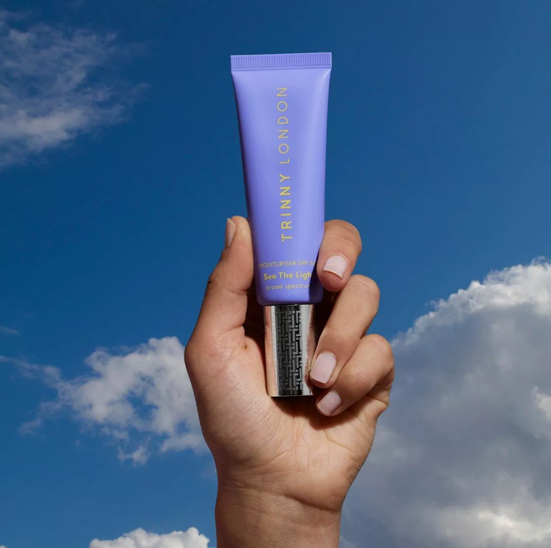 Hand holding Trinny London purple SPF moisturiser tube against blue sky with clouds, skincare product photo