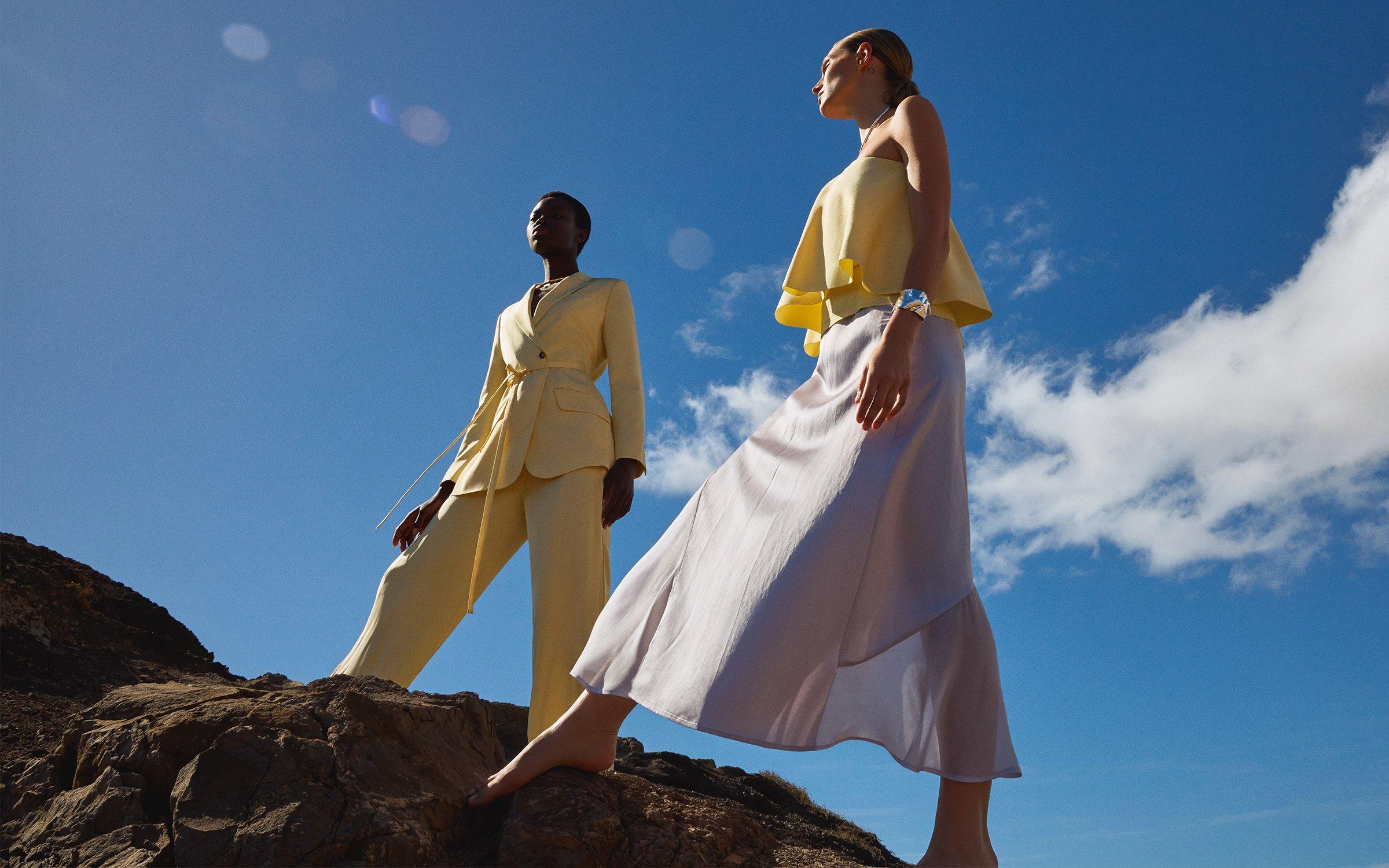 Model wearing a yellow trouser suit next to another model wearing satin skirt and top