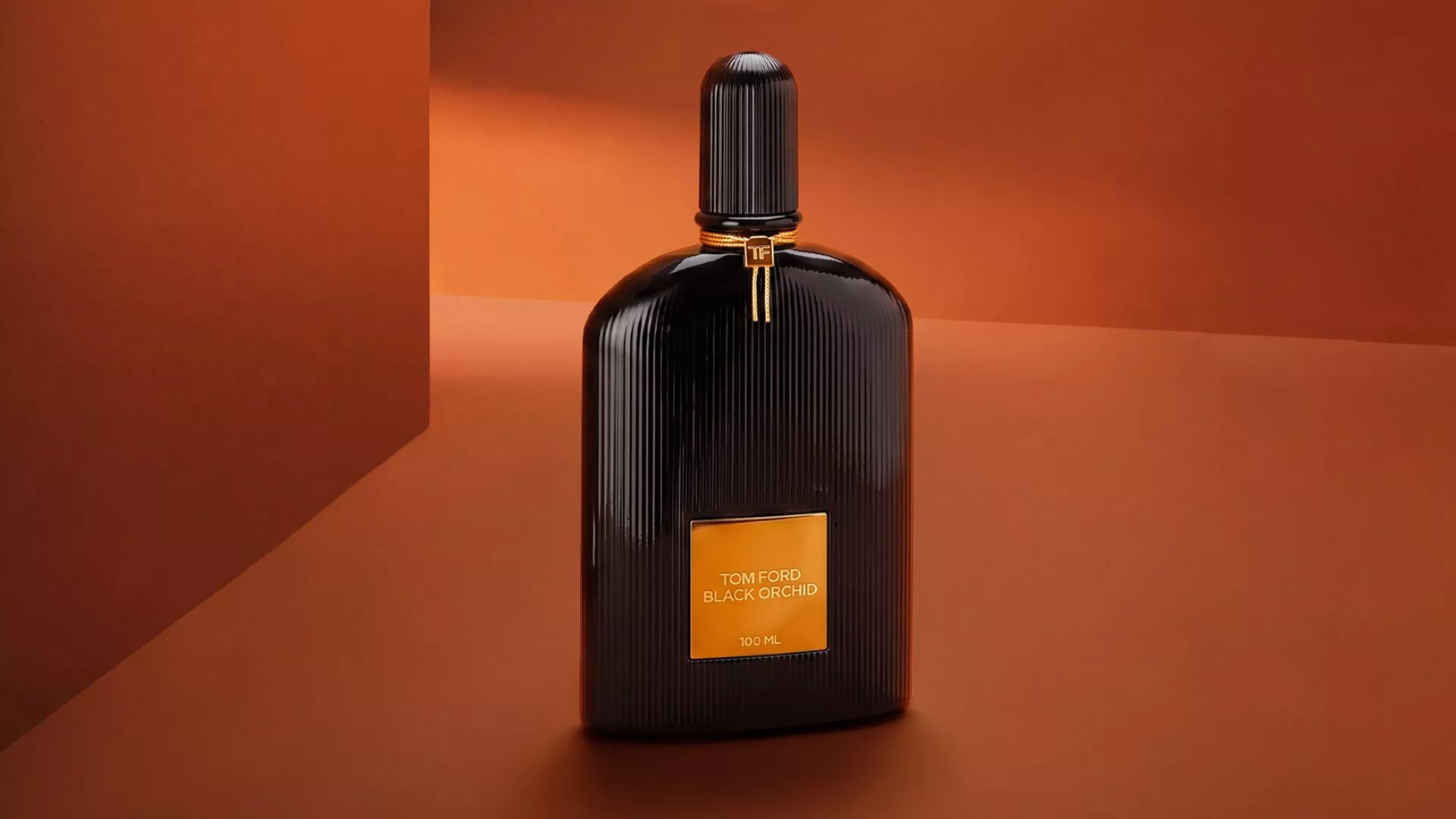 Tom Ford Black Orchid perfume bottle, black ribbed glass with gold label, on warm brown studio backdrop.