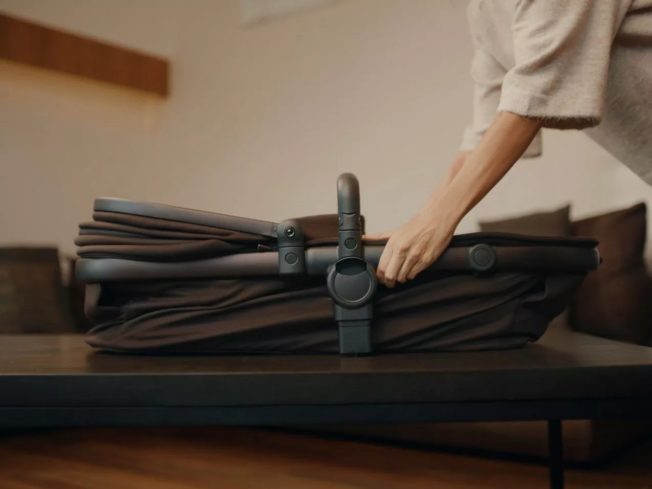 Hands folding a compact baby pram carrycot on a table in a cosy living room, travel system close-up