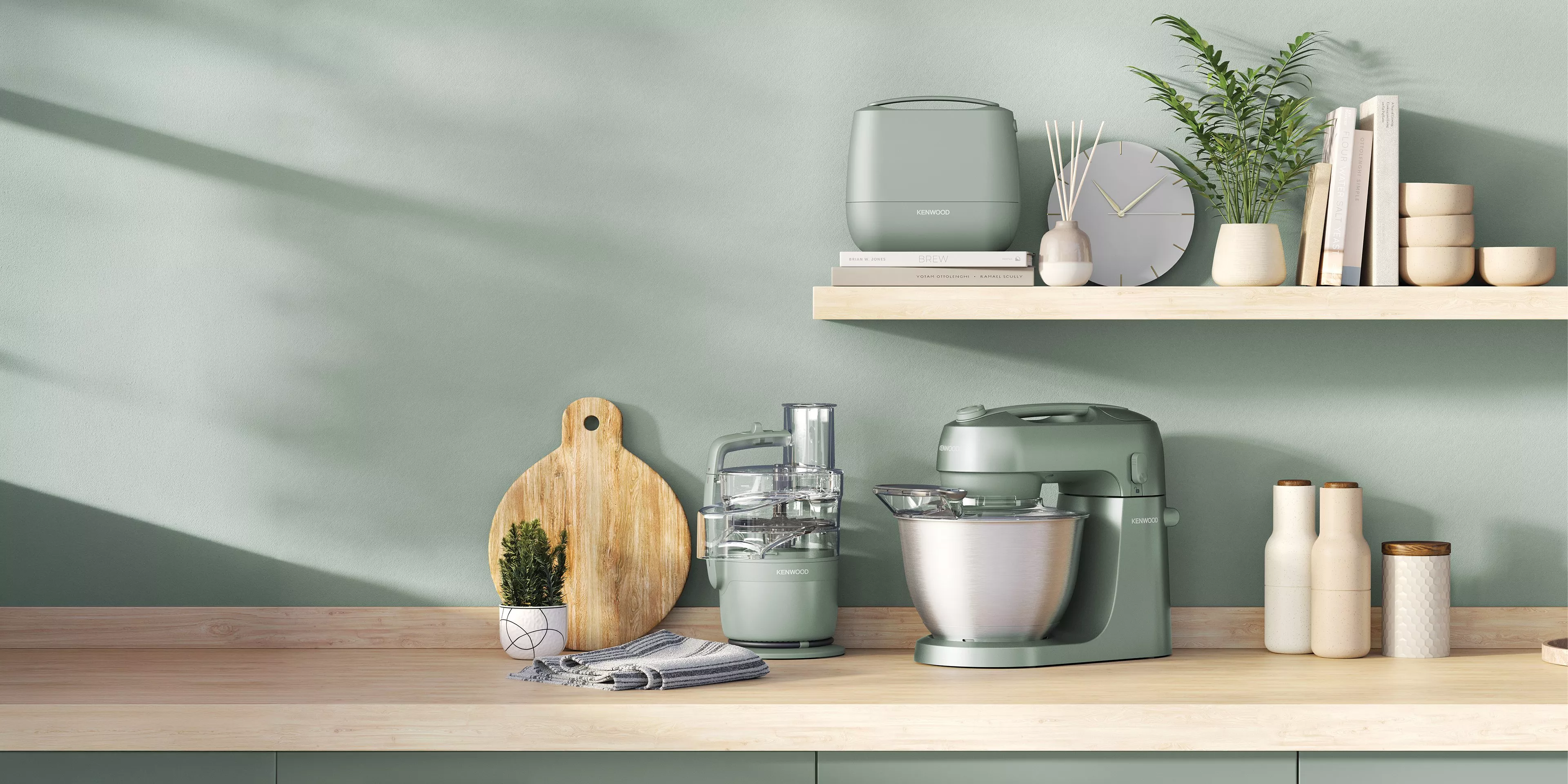 Modern sage green kitchen counter with stand mixer and food processor beneath floating shelf with plants and books