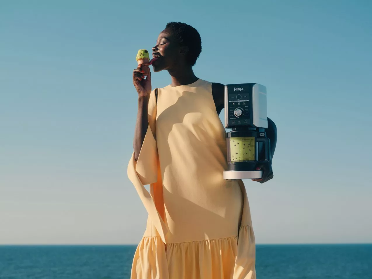 Woman enjoying an ice cream by the sea, holding a Ninja blender with green smoothie in bright sunshine