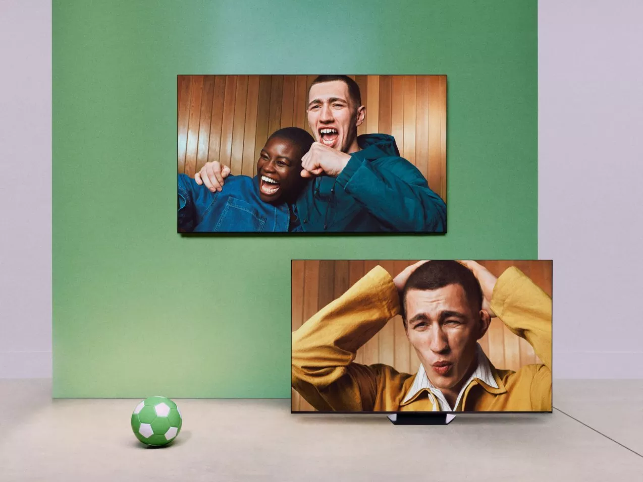 Two TVs show football fans cheering and reacting, with a green wall backdrop and a small football on floor