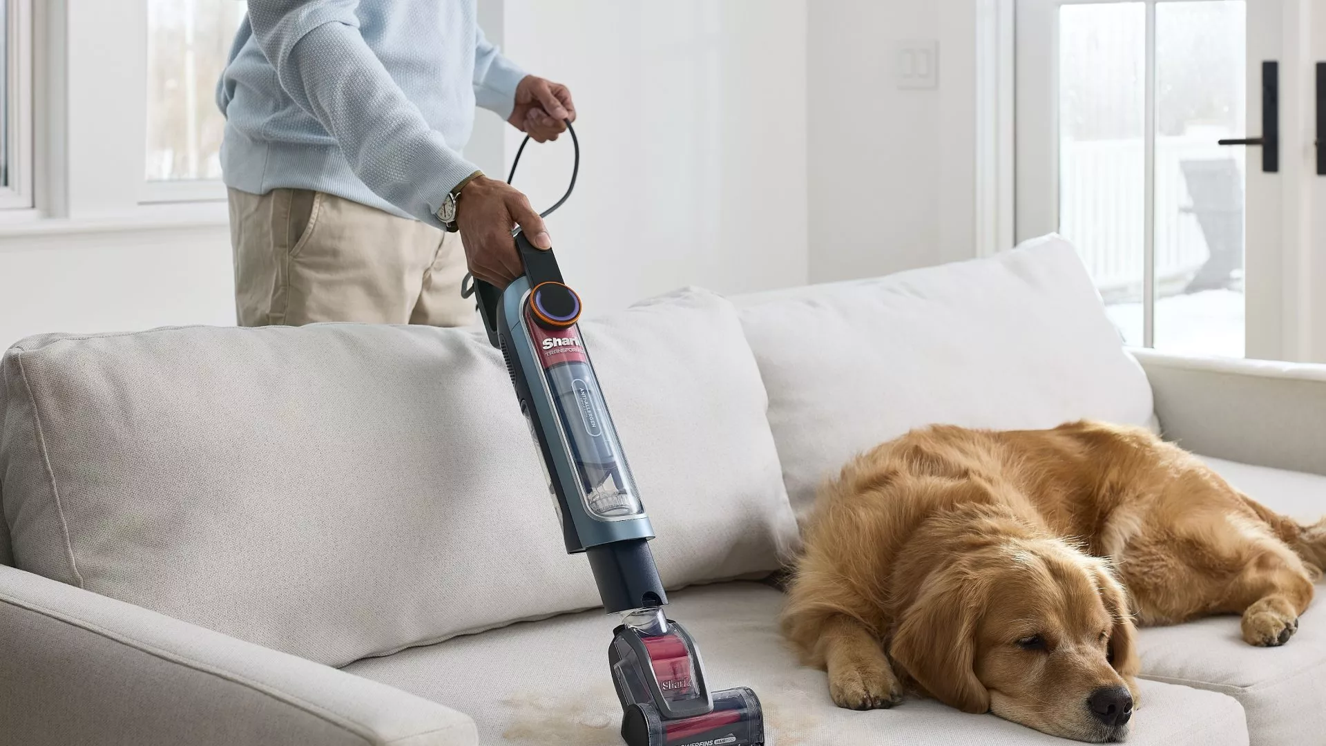 Person vacuuming a sofa beside a sleeping golden retriever, removing pet hair with a handheld cleaner indoors