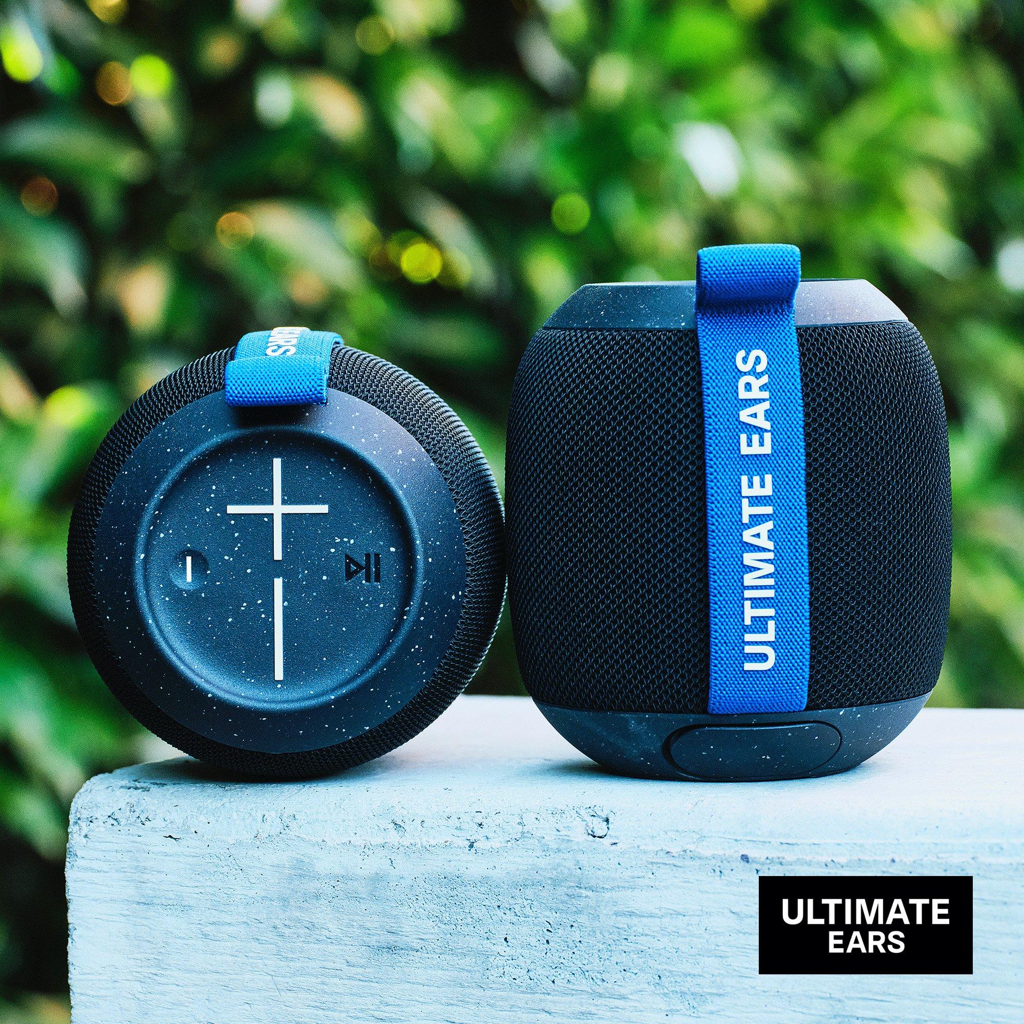 Two Ultimate Ears portable speakers with blue straps placed outdoors on a light blue surface.