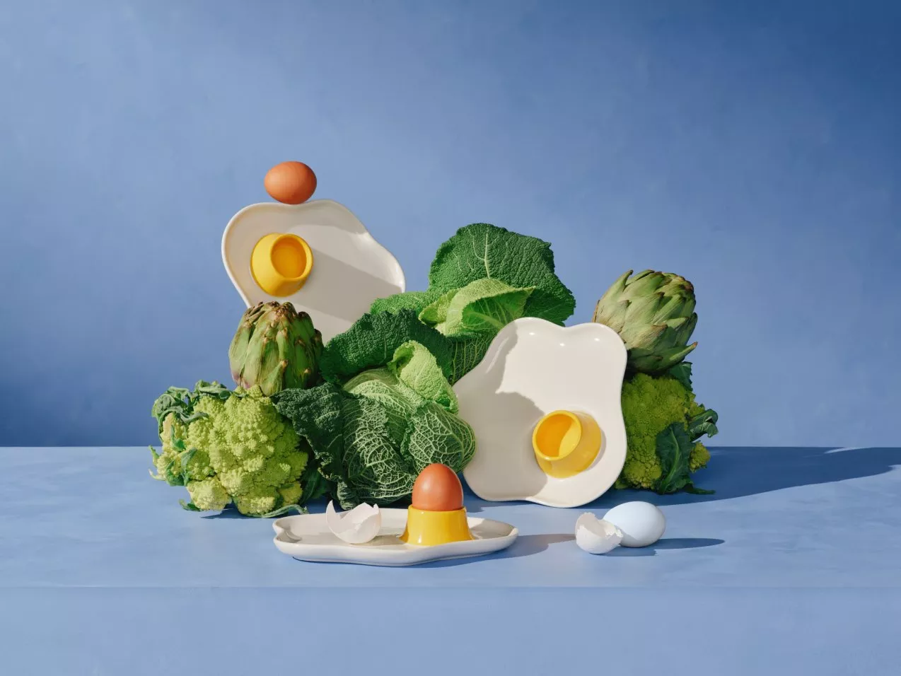Artistic arrangement with cabbage, artichokes, eggs, and abstract dishes on a blue background.