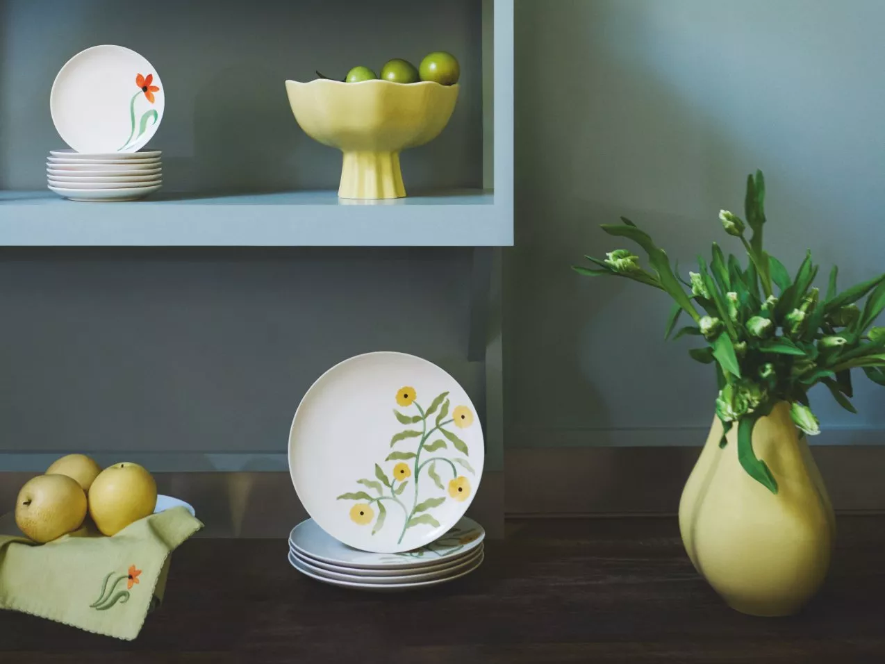 Decorative plates with floral designs, green apples in a bowl, and a vase with fresh flowers on a shelf.