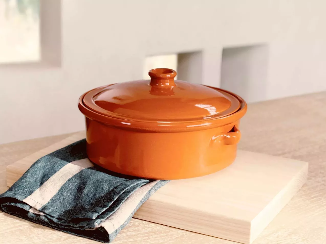 Orange glazed ceramic casserole dish with lid on wooden board, styled on kitchen table in natural light
