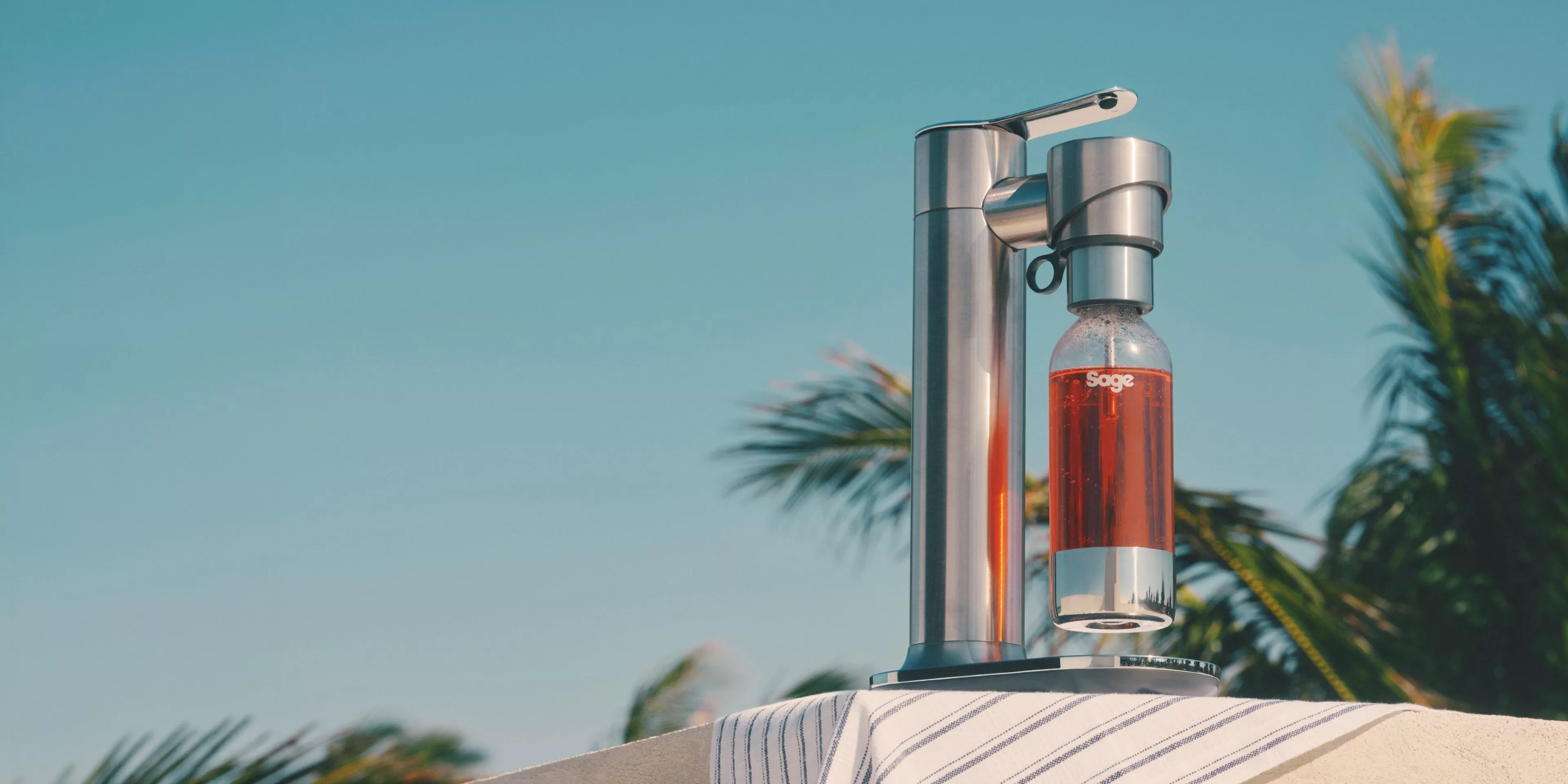 An InFizz Fusion Sparkling Drinks Maker, in Black, with blue skies and palm trees in the background