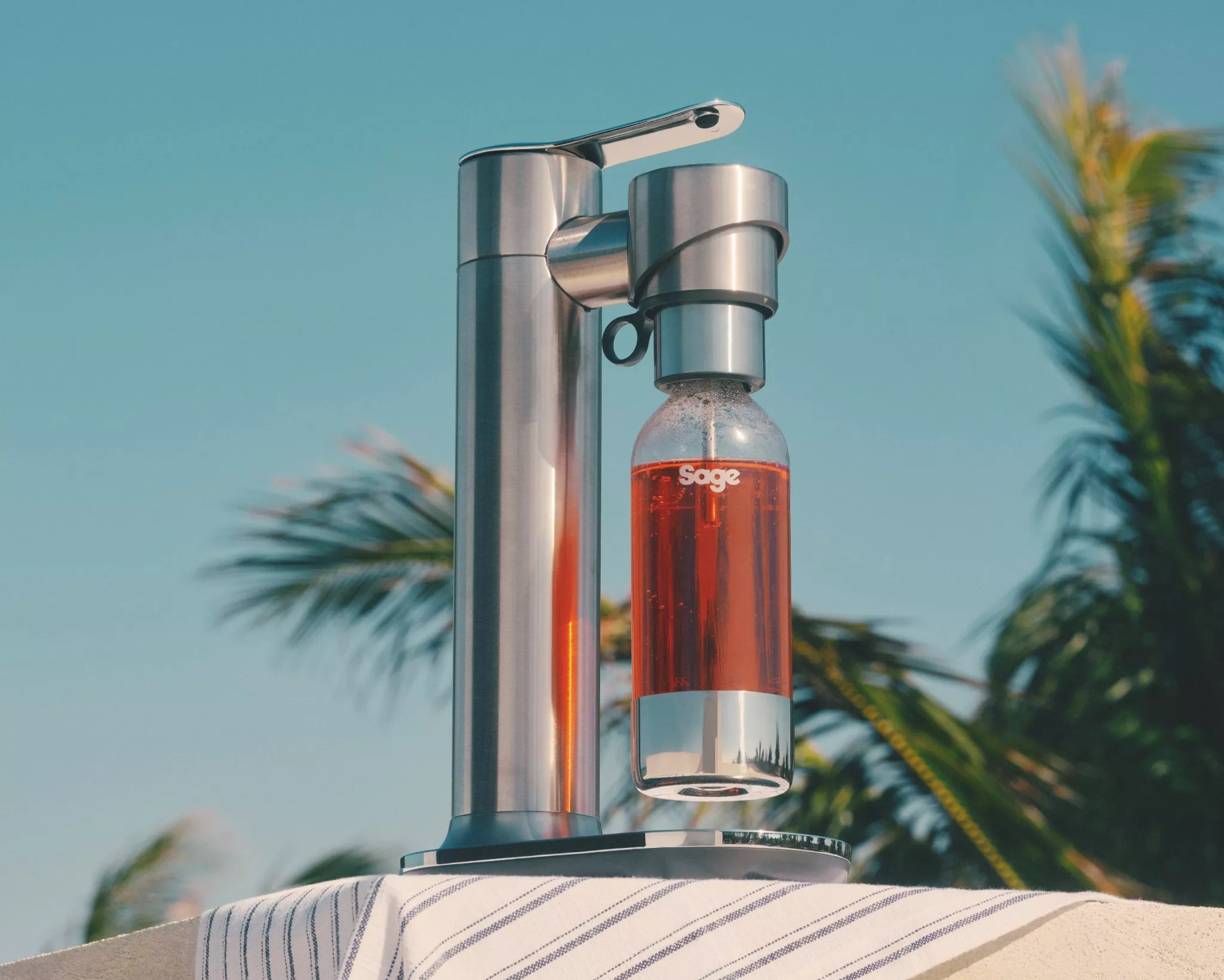 Stainless steel sparkling water maker with bottle of pink soda on outdoor table, palm trees in background