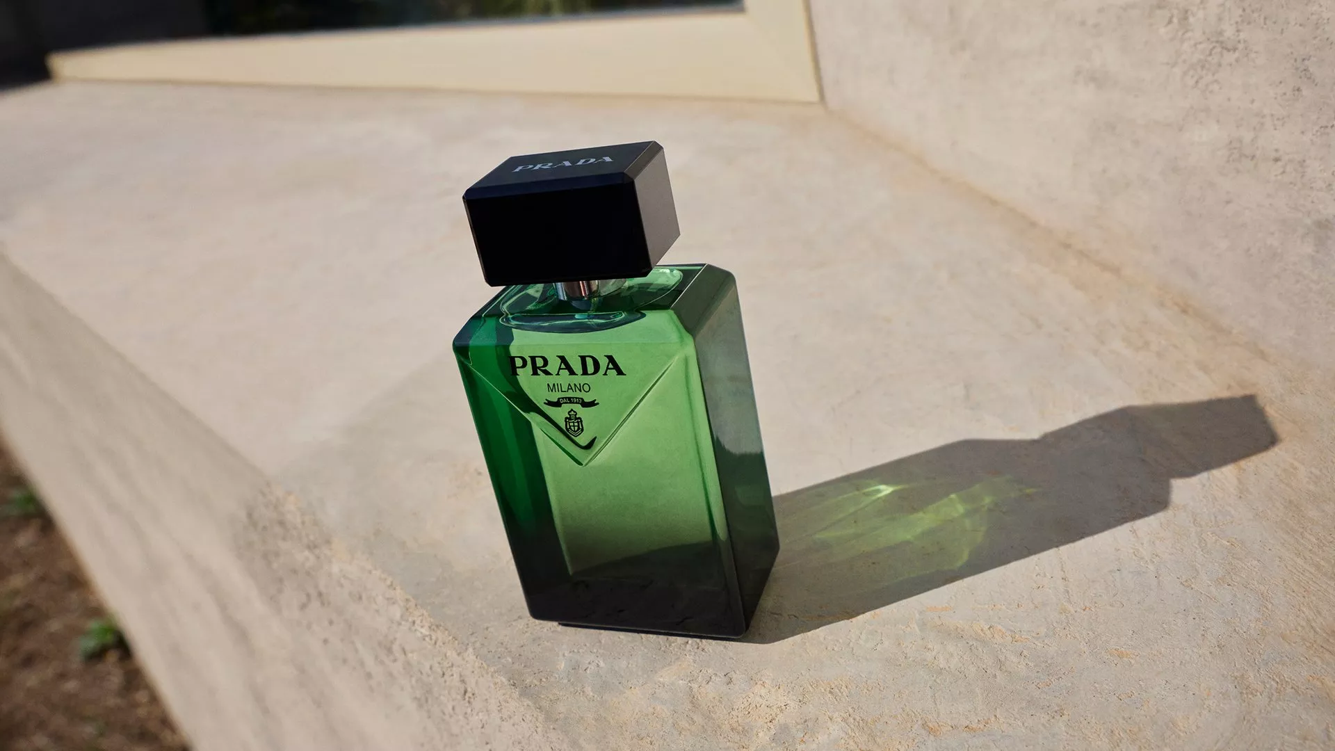 Elegant green Prada perfume bottle with black cap casts a shadow on a sunlit beige surface. Modern luxury design.