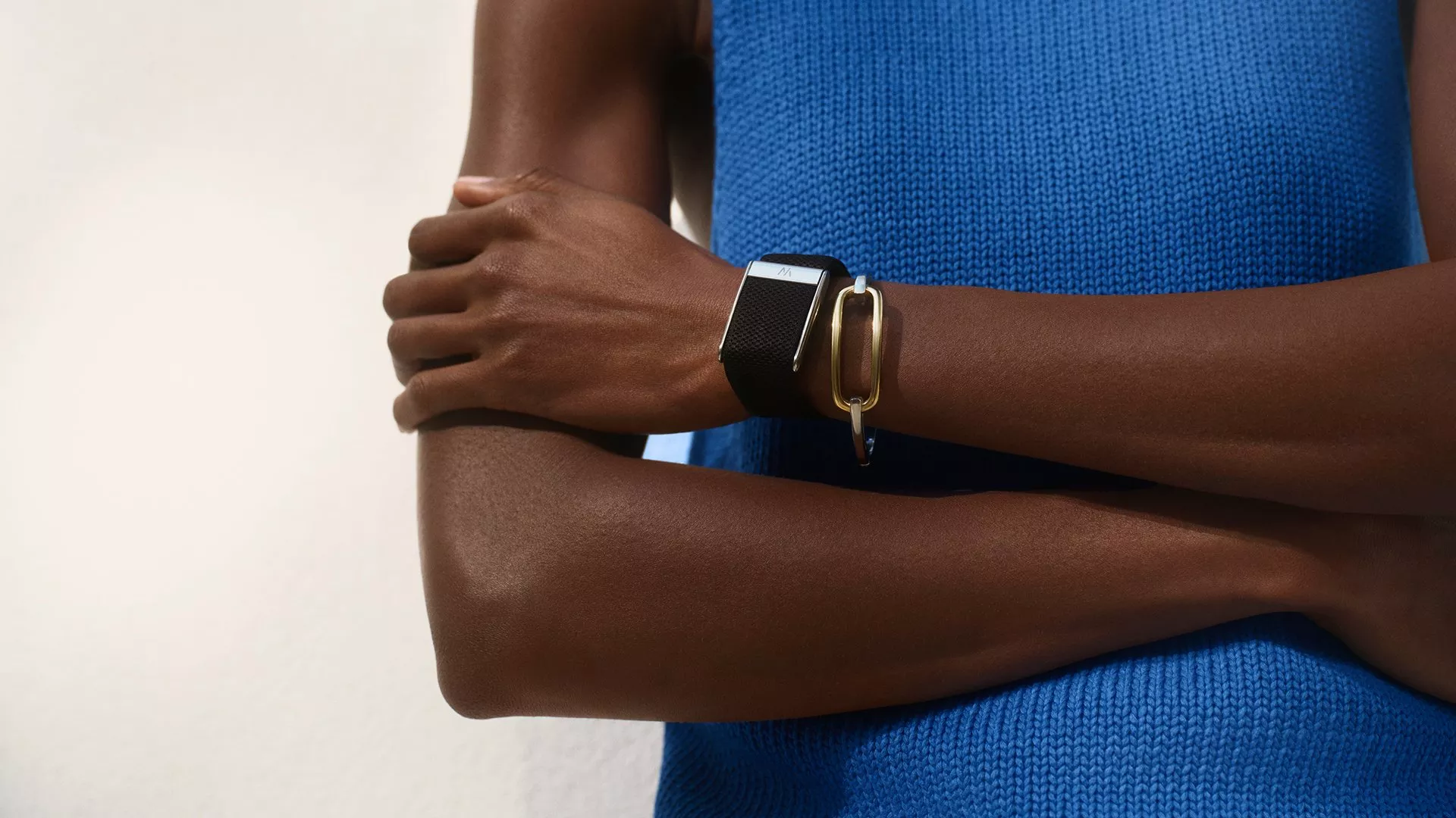 Person wearing a blue top with arms crossed, showcasing a black smartwatch and a gold bracelet.