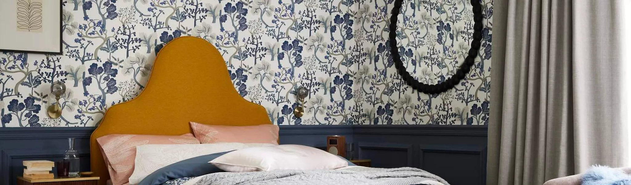 Stylish bedroom with ochre upholstered headboard, blue floral wallpaper, navy panelling and grey curtains.