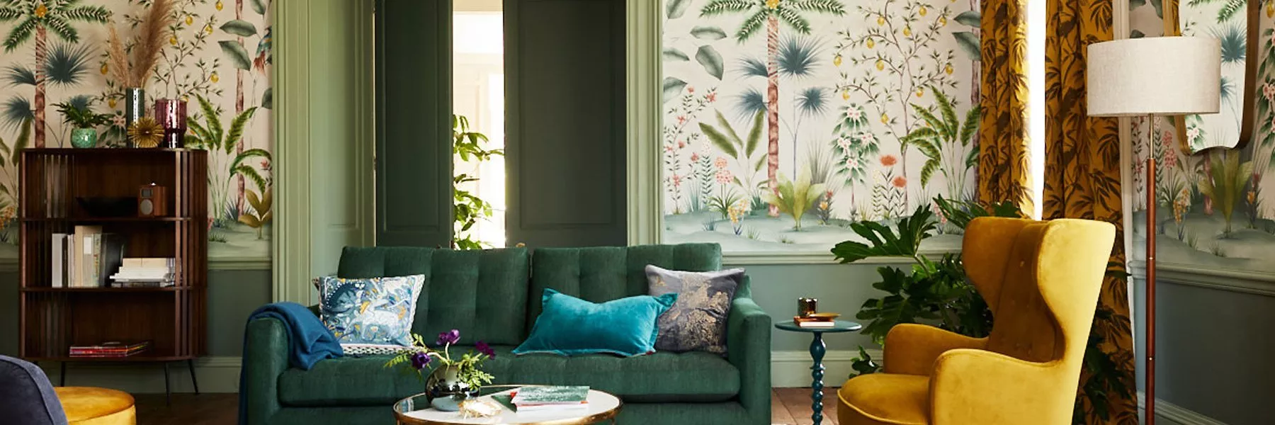 Stylish living room with green velvet sofa, mustard armchair, botanical wallpaper, plants and warm lighting decor