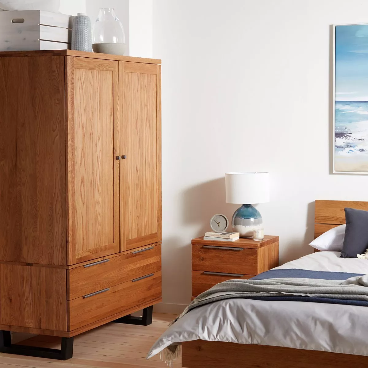 Bedroom with wooden wardrobe, bedside table, and bed. Lamp, clock, and decor items on the table. Coastal artwork.