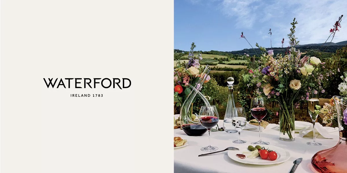 An image showing an outdoor dining table in a vineyard, set with many wine glasses, bottles, and floral arrangements, featuring a long, white tablecloth.
