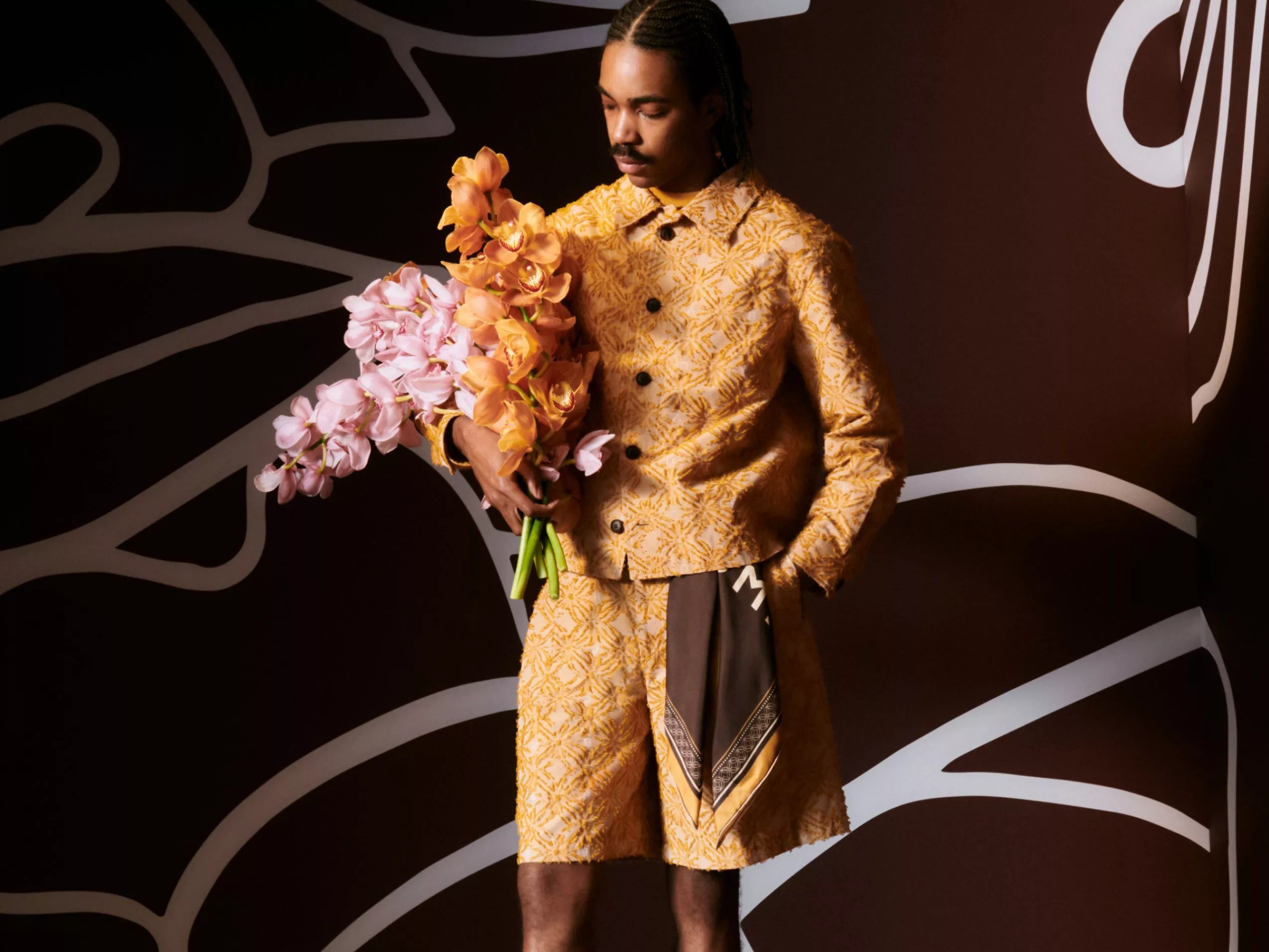 Man in a patterned yellow outfit holding pink and orange flowers against a dark, abstract background.