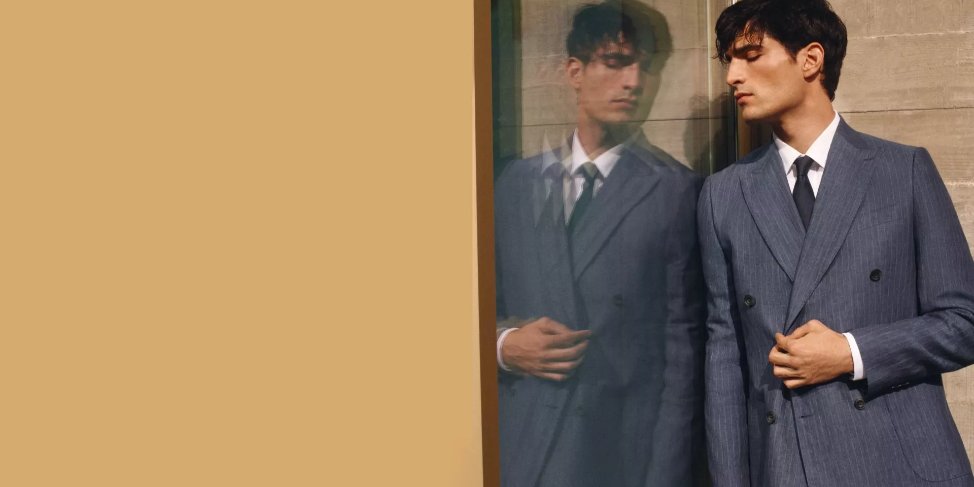 Man in a blue pinstripe suit stands by a reflective glass, showcasing elegant formal wear against a beige backdrop.