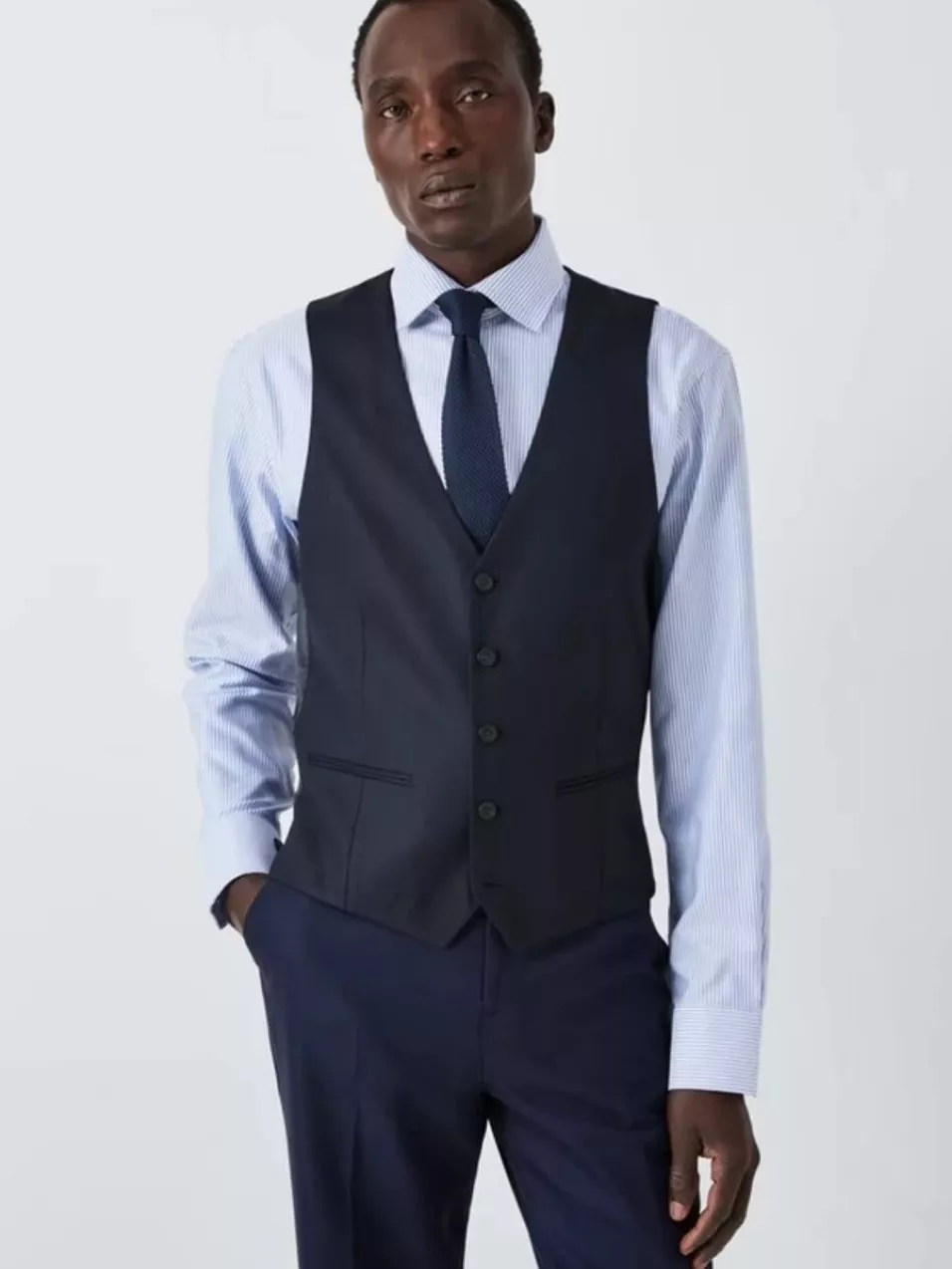 A man in a stylish navy waistcoat and trousers, paired with a light blue striped shirt and a dark tie.