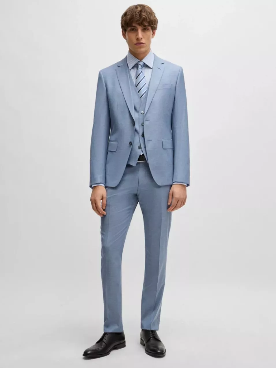 Man in a tailored light blue suit with matching waistcoat, striped shirt and tie, wearing black shoes.