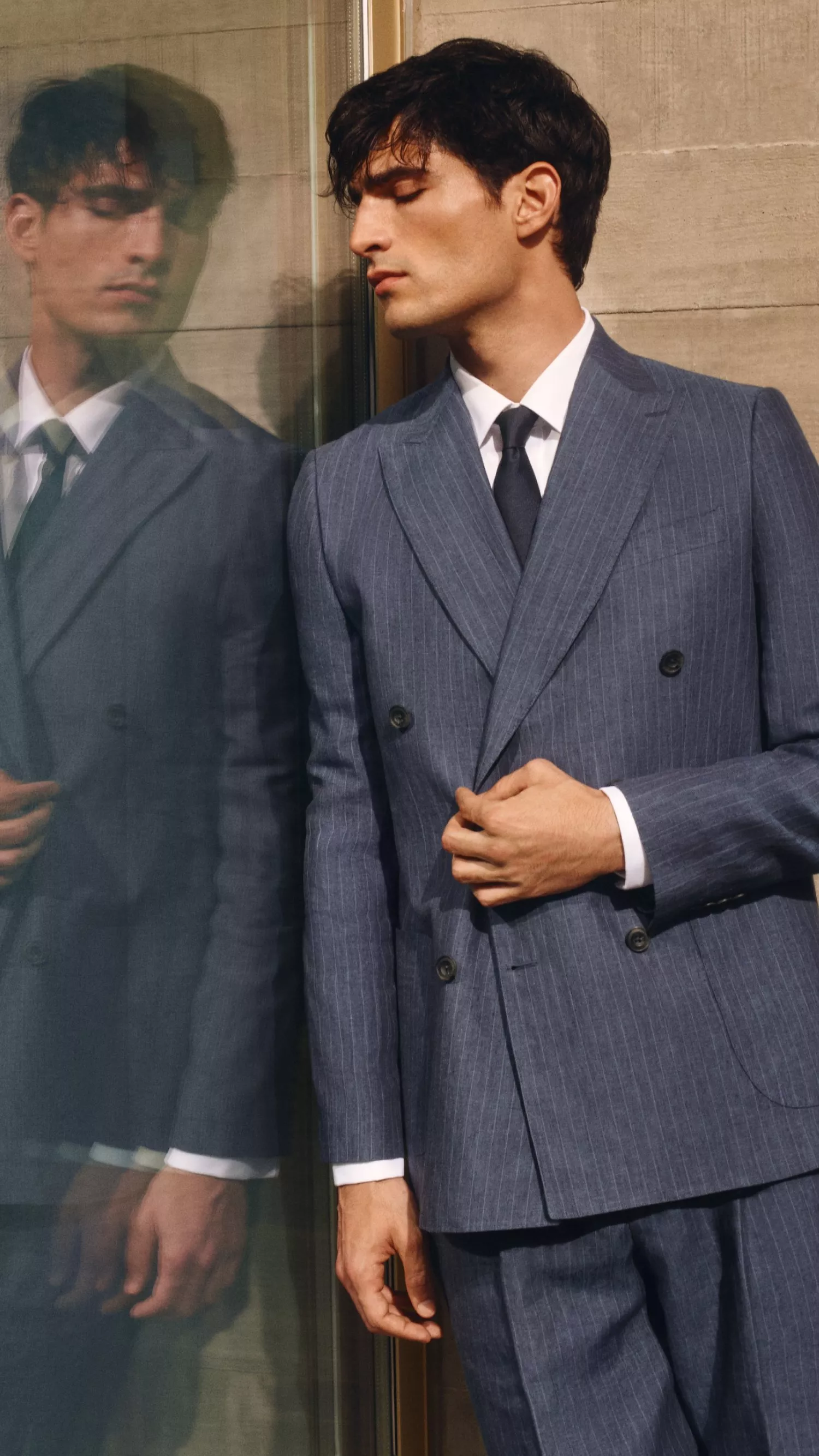 Man in tailored blue pinstripe double-breasted suit and tie, leaning by glass wall, reflecting modern formal menswear style