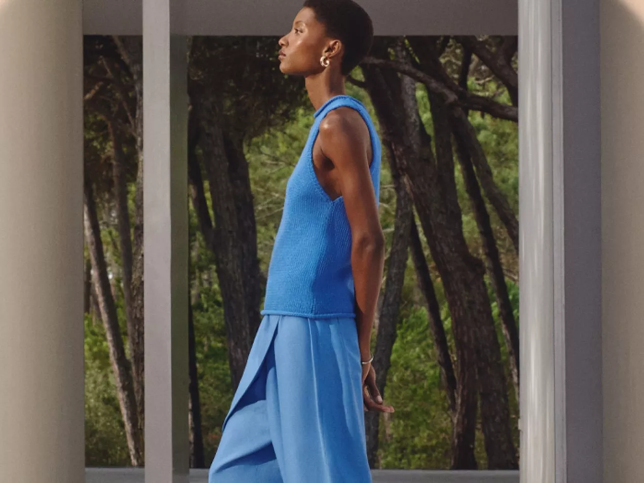 A person in a sleeveless blue top and skirt walks through a modern architectural space; trees in the background.