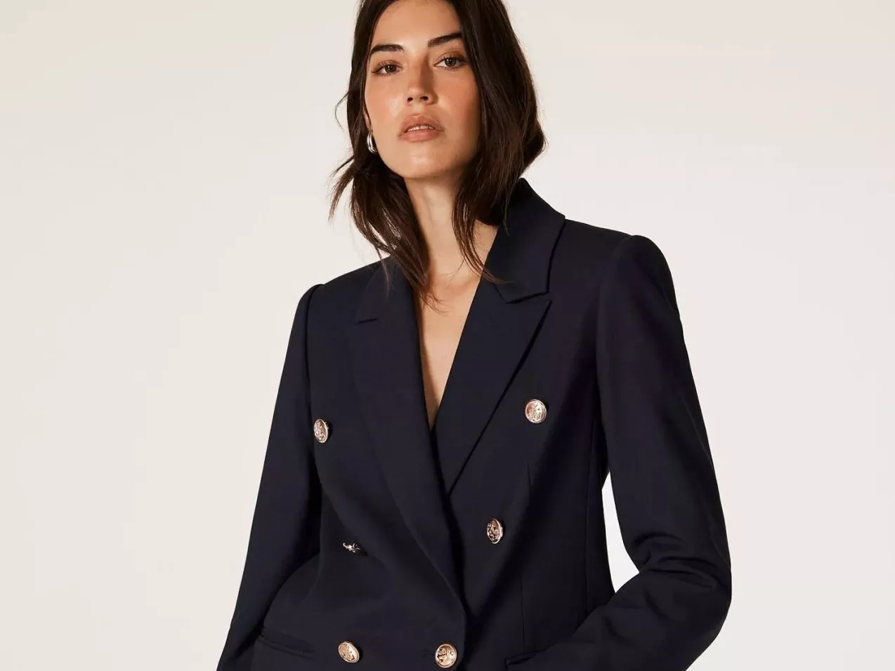 Woman in a dark double-breasted blazer with gold buttons, posing against a neutral background.