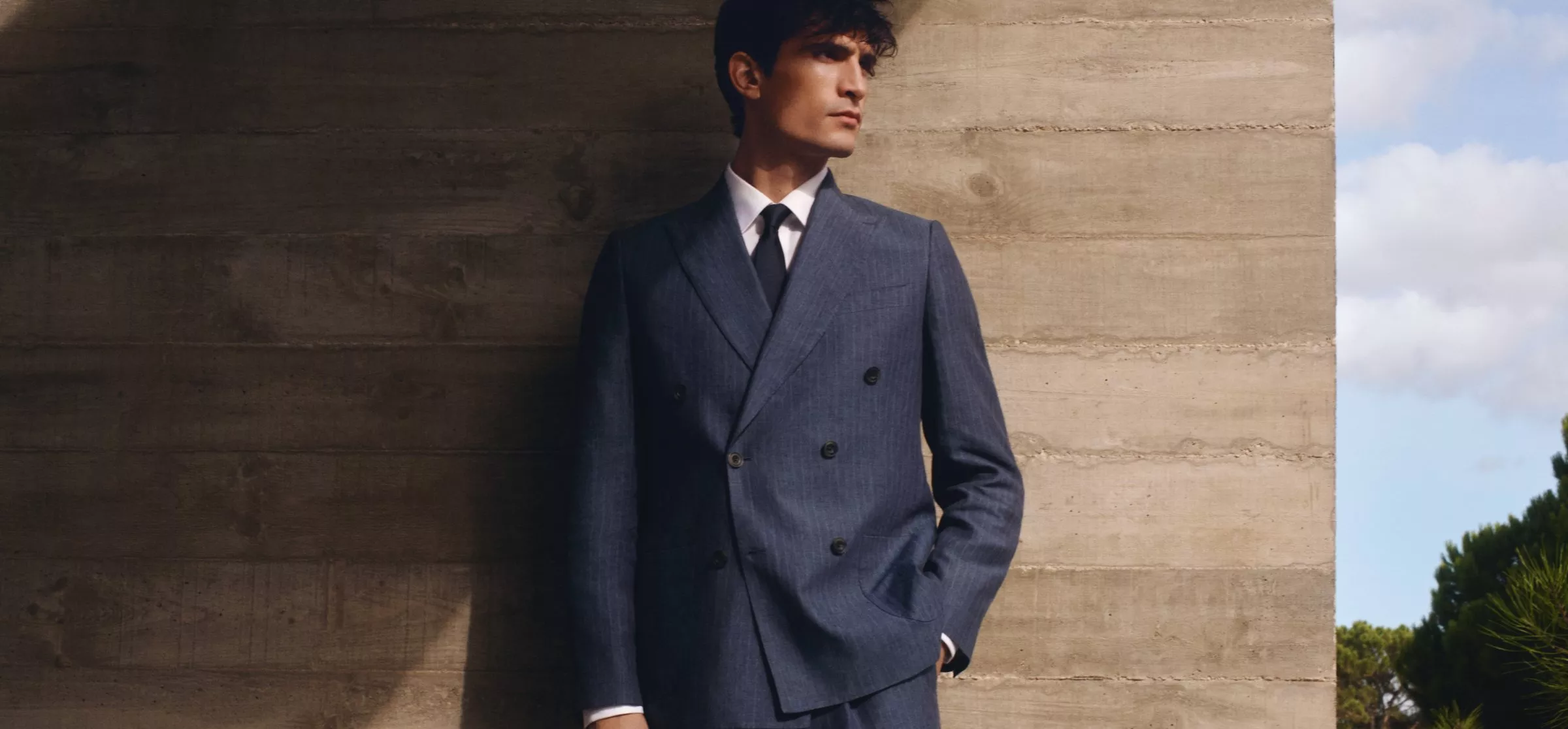 A man in a stylish double-breasted blue suit leans against a textured concrete wall outdoors.