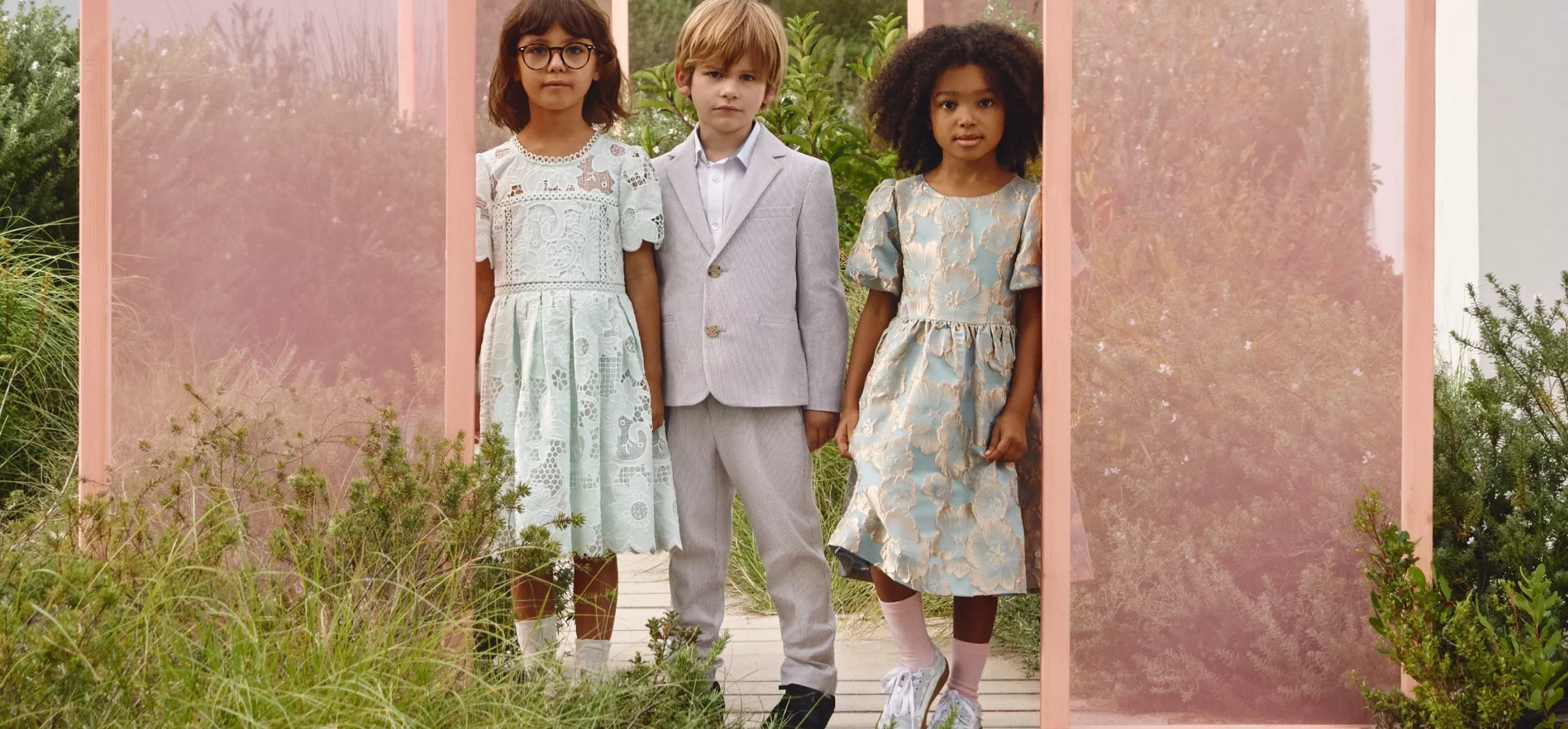 Three children in elegant outfits stand outdoors against a backdrop of greenery and pink panels.