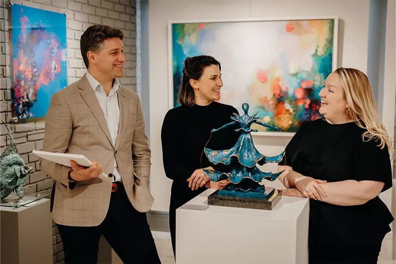 Art gallery staff discussing a blue abstract sculpture beside colourful modern paintings in a contemporary showroom.