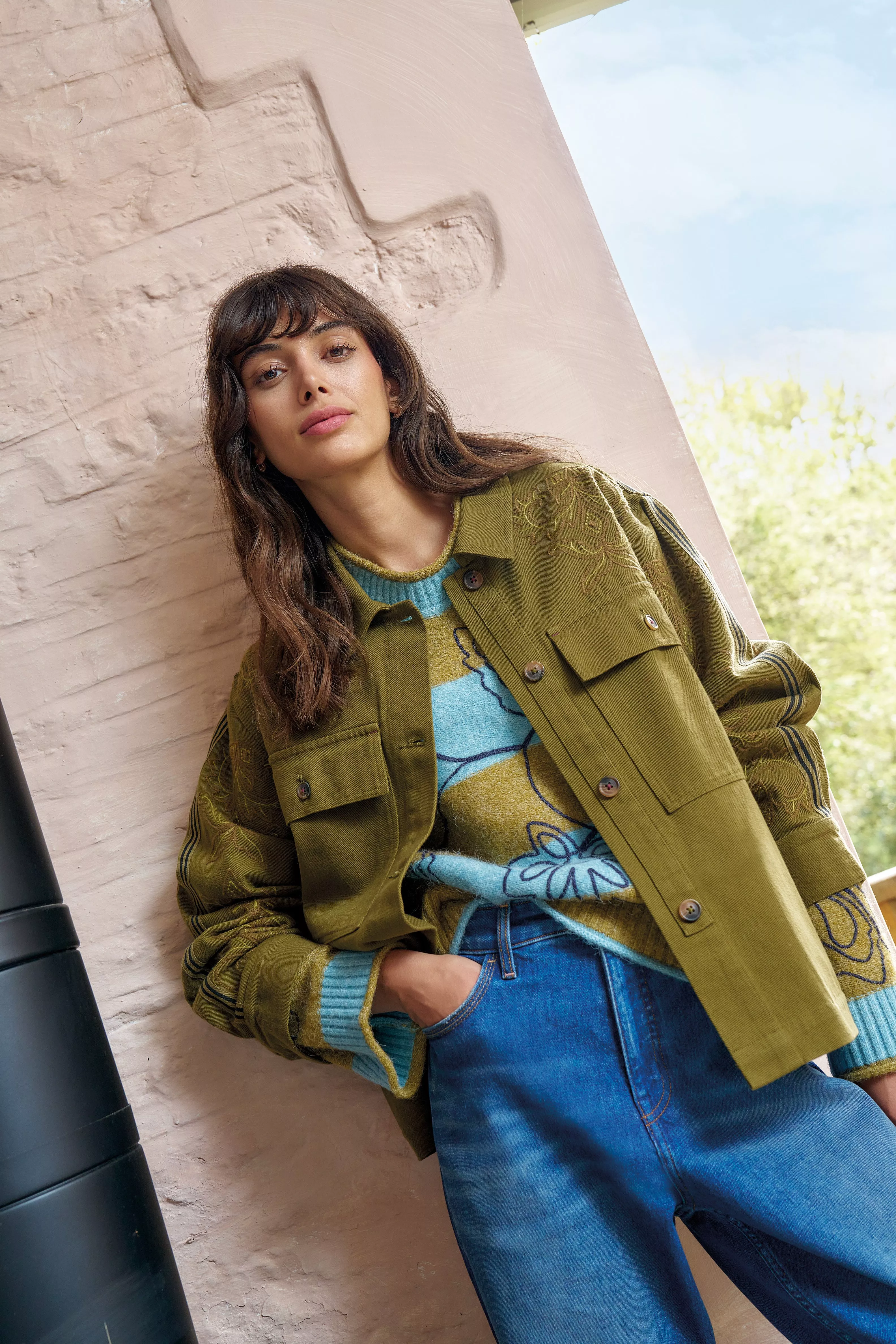 Fashion portrait of a woman in olive jacket, floral jumper and blue jeans, leaning against a stone wall outdoors